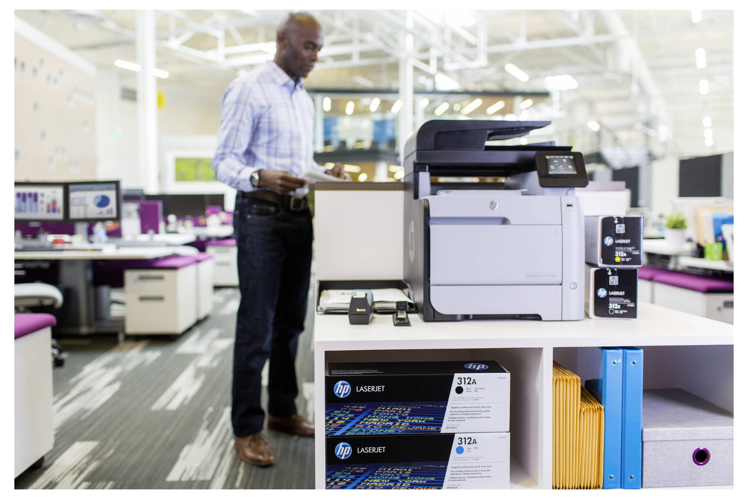 Een man staat naast een HP LaserJet-printer op een showroom-displaytafel in een kantooromgeving, met tonercartridges en voorraden zichtbaar eronder.