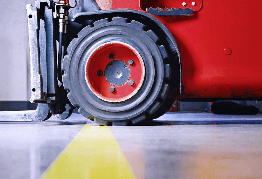 Een rode heftruck rijdt over een gele lijn op een betonnen vloer in een opslagloods. Alleen het voorwiel is zichtbaar.