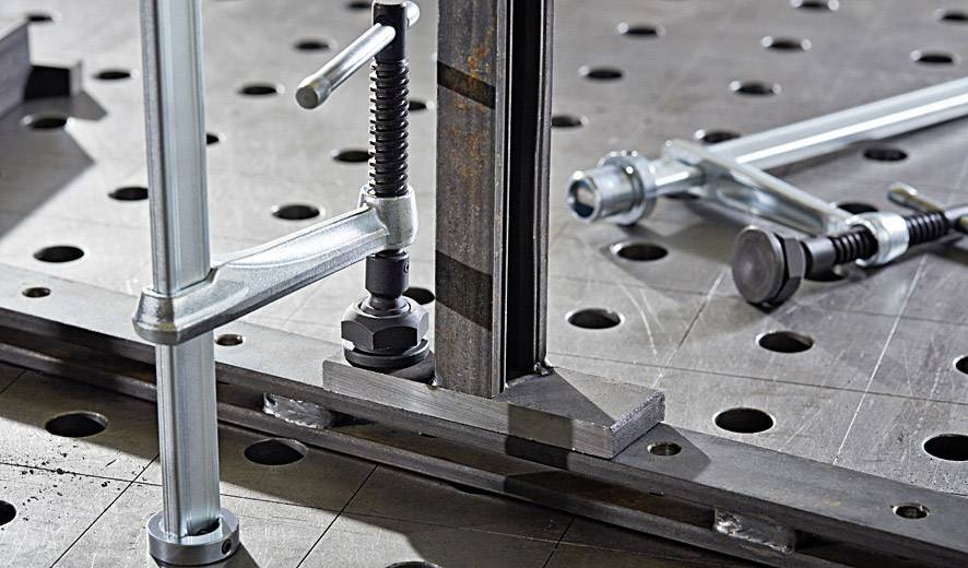 Metalen klemmen en schroeven bevestigen een stalen plaat met gatenpatroon op een werktafel, weergegeven in close-up.