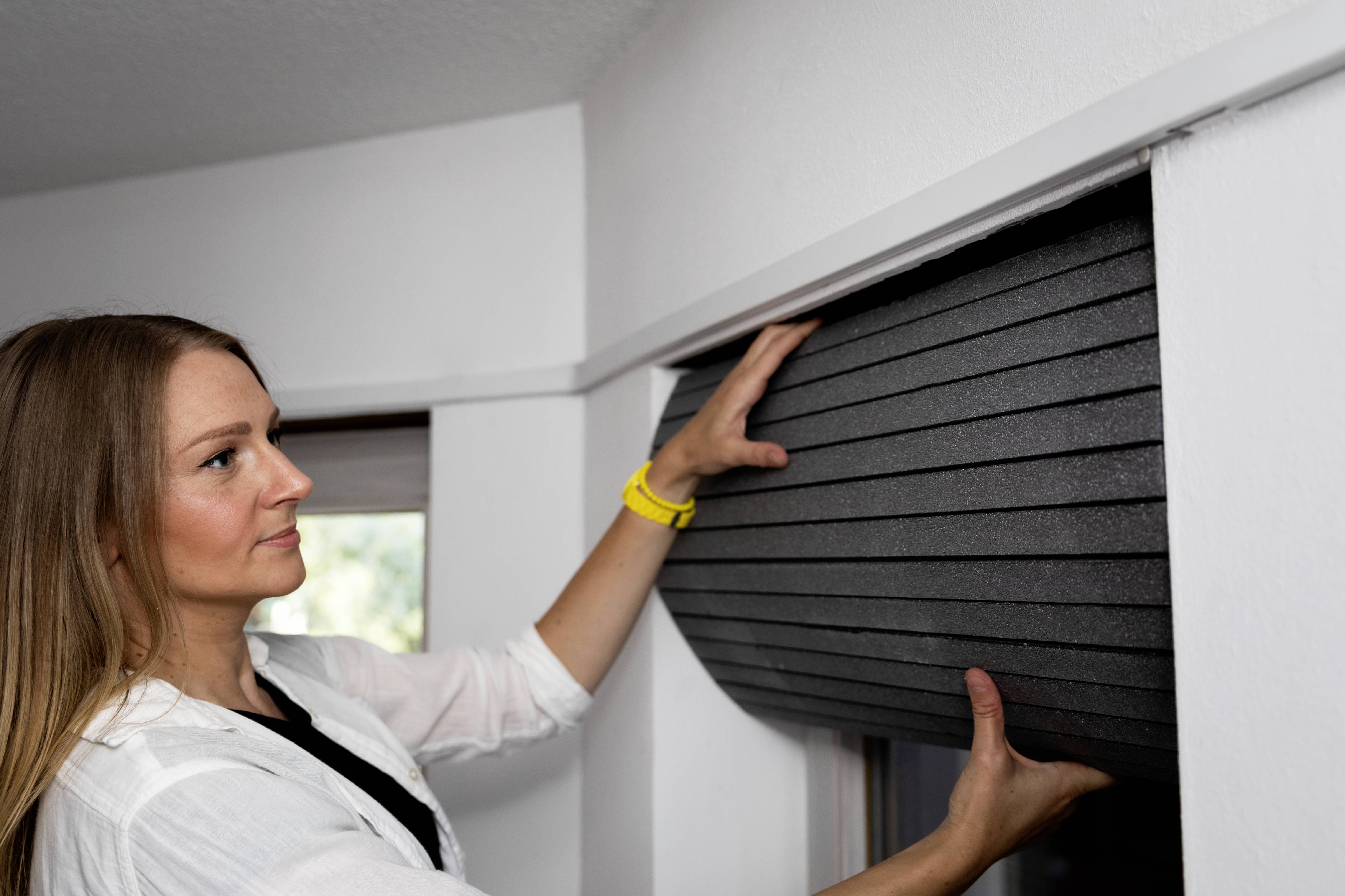 Een vrouw tilt een zwarte rolluik in een kamer op. Ze draagt een wit overhemd en een geel armbandje.