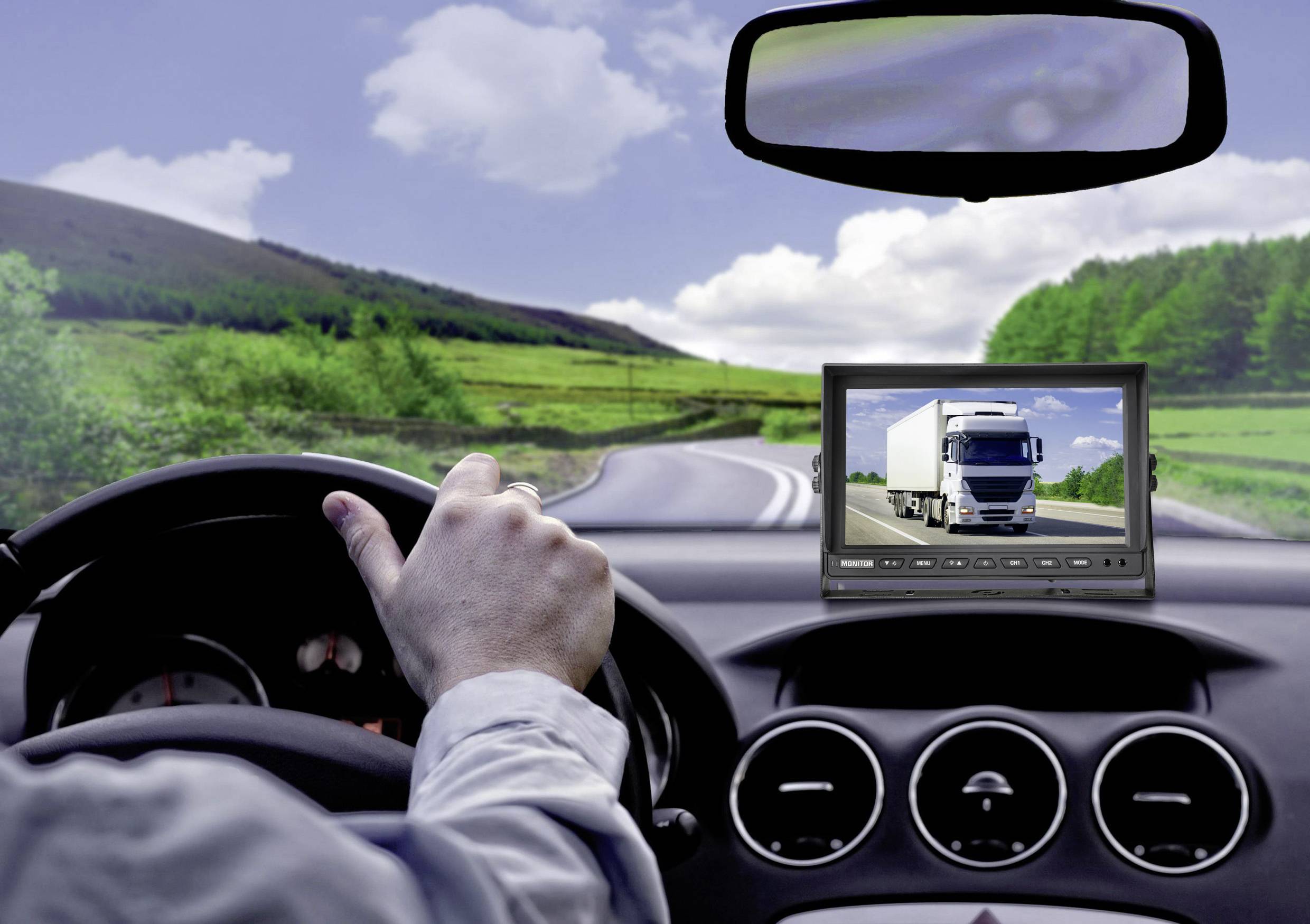 Een bestuurder houdt het stuur van een auto vast. Op een monitor is een grote vrachtwagen te zien. De weg loopt door een groene landschap.