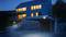 Modern huis met grote ramen en blauwe gevelverlichting 's nachts, met stenen trap en een carport op de voorgrond.