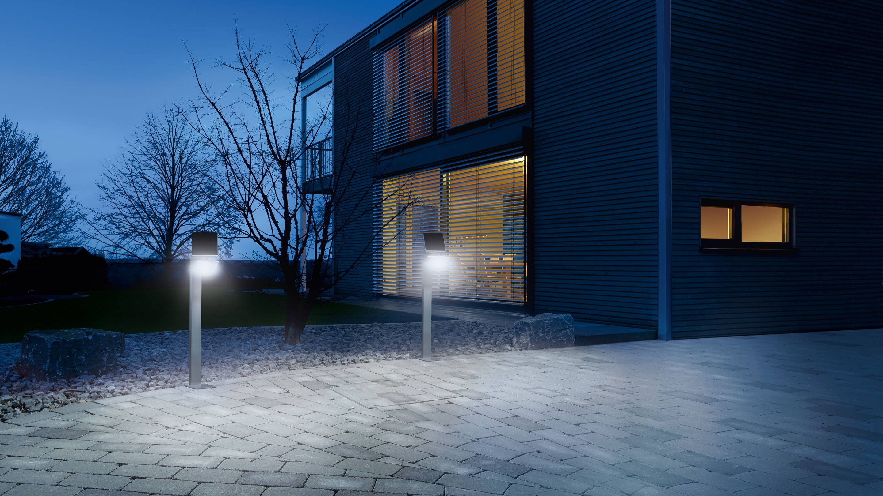 Een modern huis bij schemering, verlicht door buitenlampen. Ramen stralen warm licht uit, een kale boom op de achtergrond.