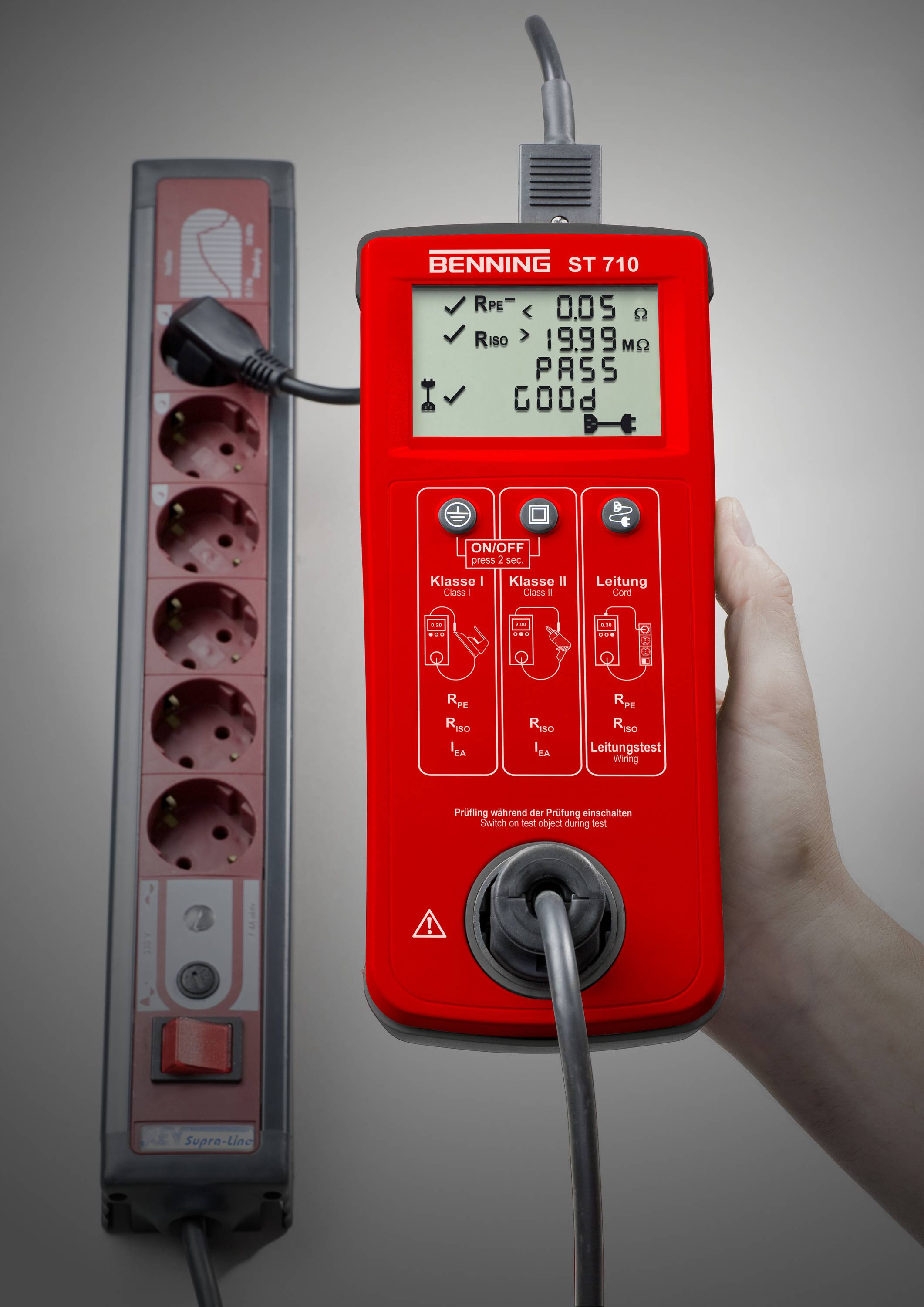 Een rode elektrische tester wordt vastgehouden door een hand, met 'PASS' op het scherm. Naast de tester staat een stekkerdoos.