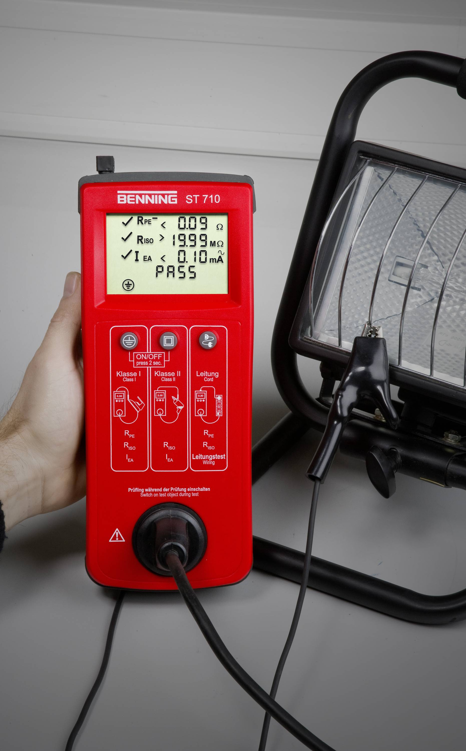 Een handapparaat test de veiligheid van een werklamp. Het display toont weerstandswaarden en een 'PASS'-status.