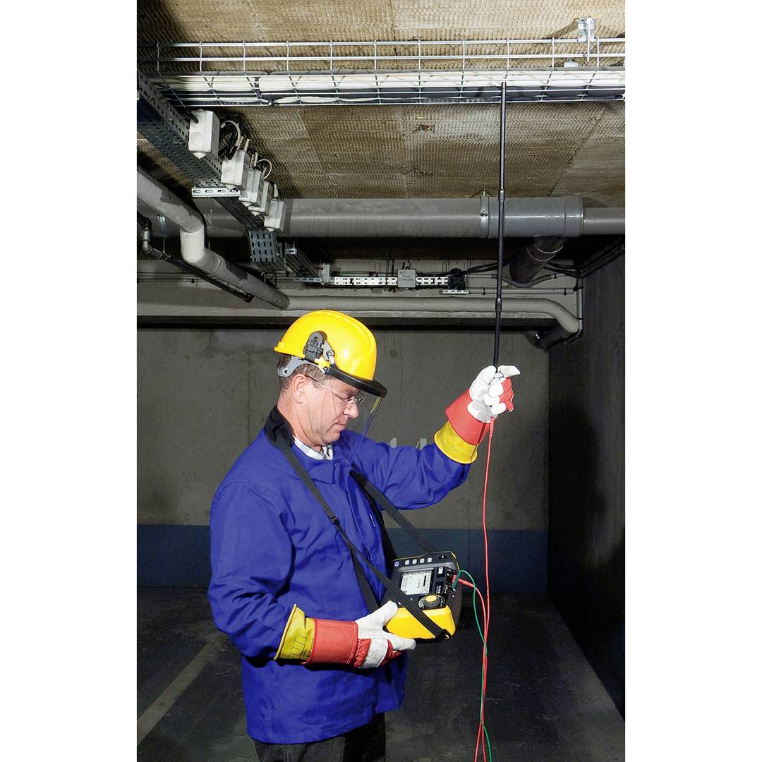 Een man in werkkleding en veiligheidshelm meet met een apparaat de elektrische spanning in een kabel in een industrieel gebouw.