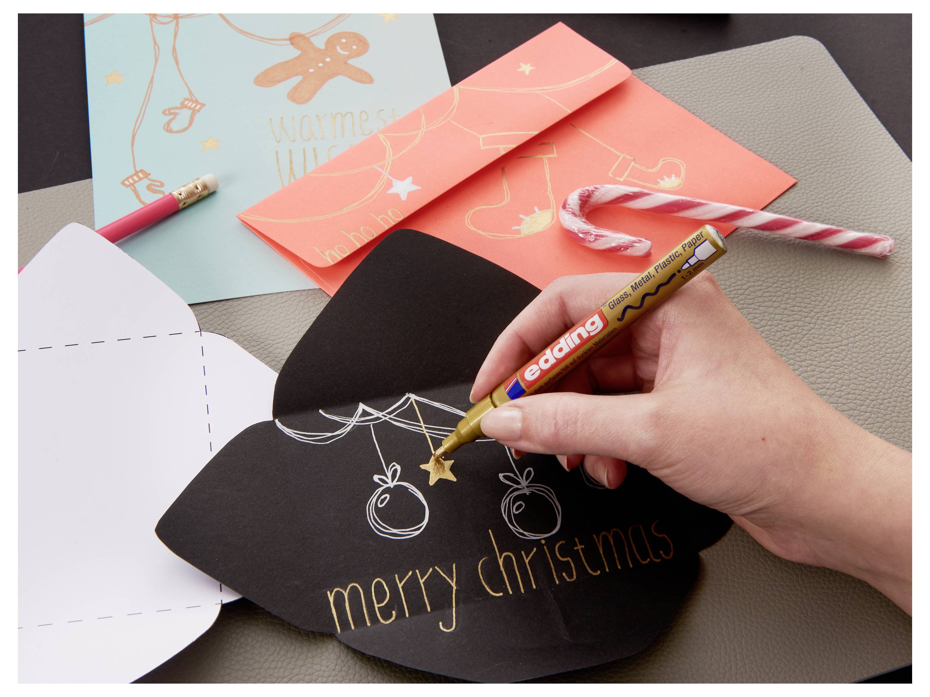 Handschrift 'fijne kerst' met een goudkleurige marker op een zwart gevouwen kaart, omgeven door kleurrijke handgemaakte vakantiekaarten en pennen.