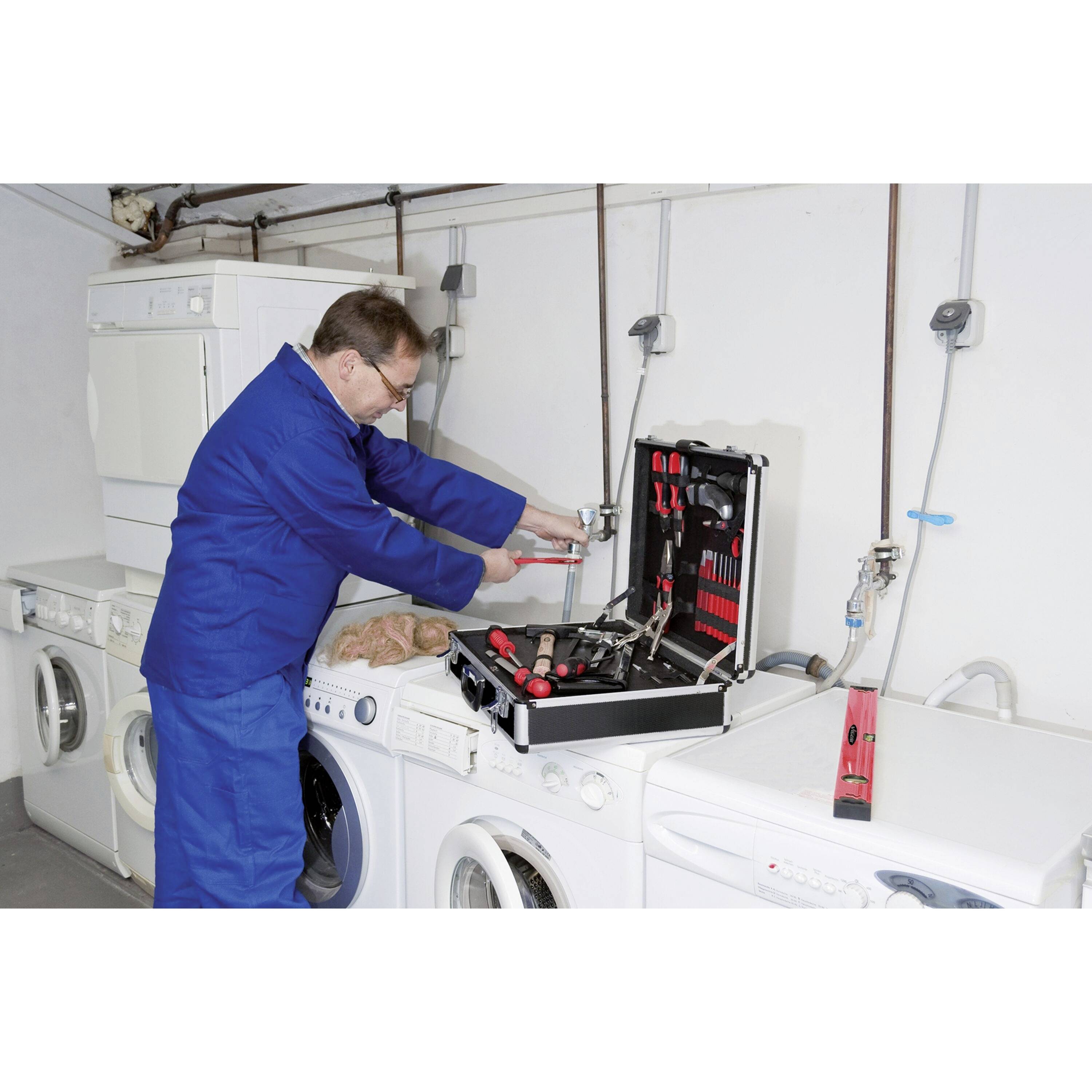 Een technicus in een blauwe overall gebruikt gereedschappen uit zijn kit om een wasmachine te repareren in een wasruimte. Verschillende machines staan langs de muur.