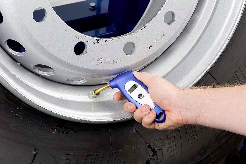 Een hand gebruikt een digitale manometer om de bandenspanning van een band aan een zilveren velg te controleren.