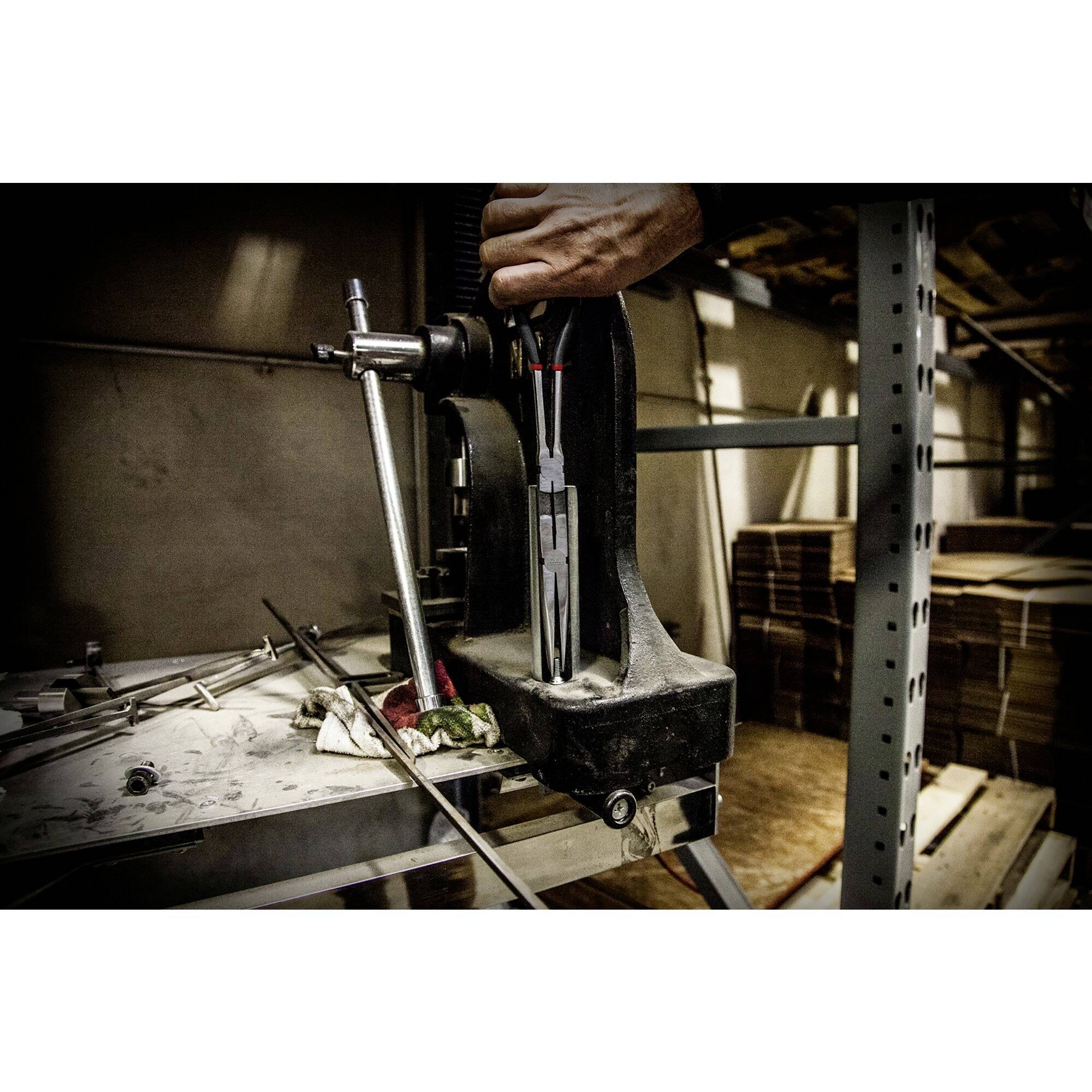 Een bankschroef op een werkbank in een werkplaats, bediend door een hand. Op de achtergrond zijn planken met materialen zichtbaar.