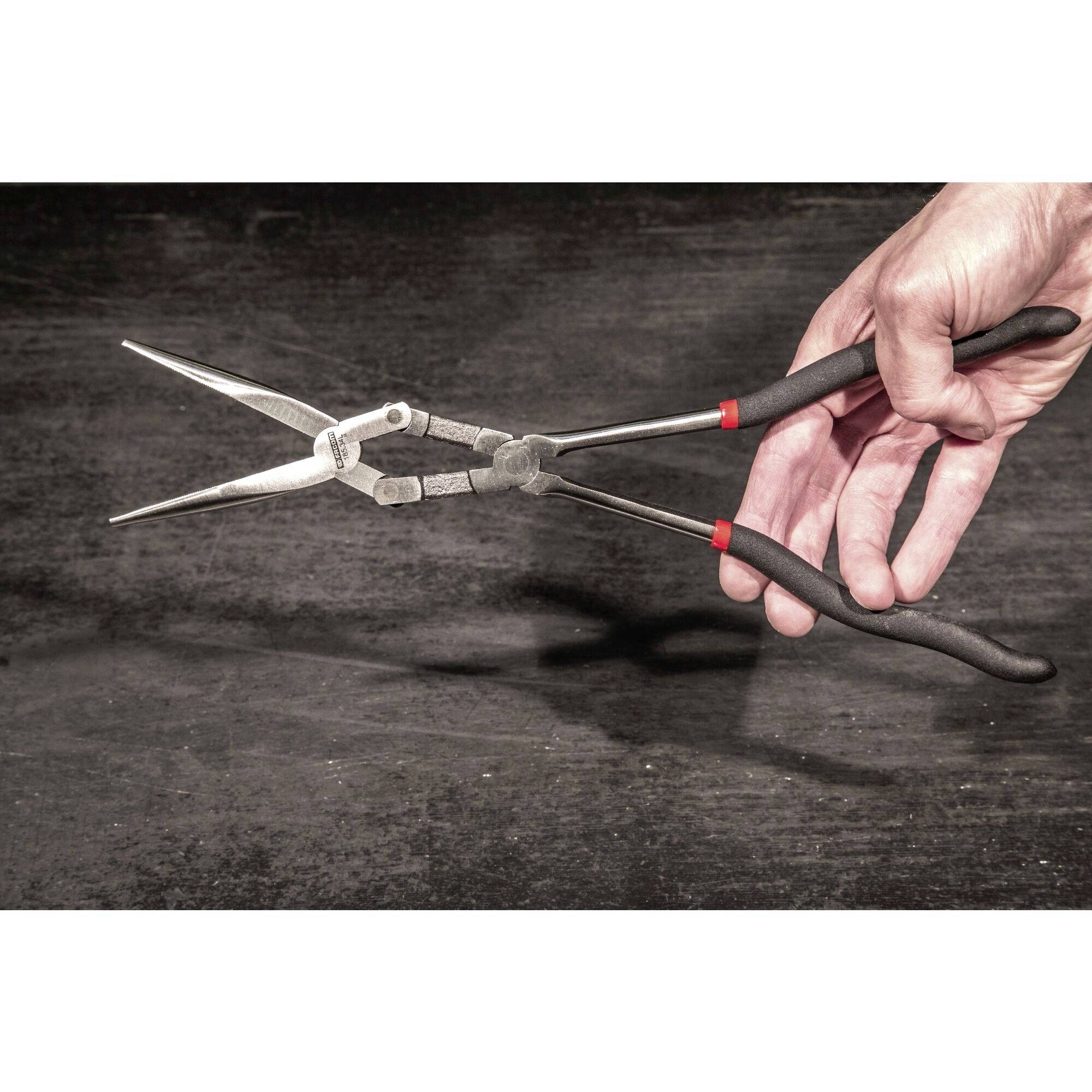 Een hand houdt een zwarte tang vast boven een donkere achtergrond. De tang is lang en heeft rode markeringen op de handgreep.