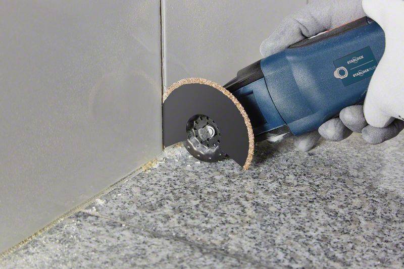 'Een hand houdt een elektrisch multifunctioneel gereedschap met een schuurblad dat werkt langs de rand van tegels en vloer.'