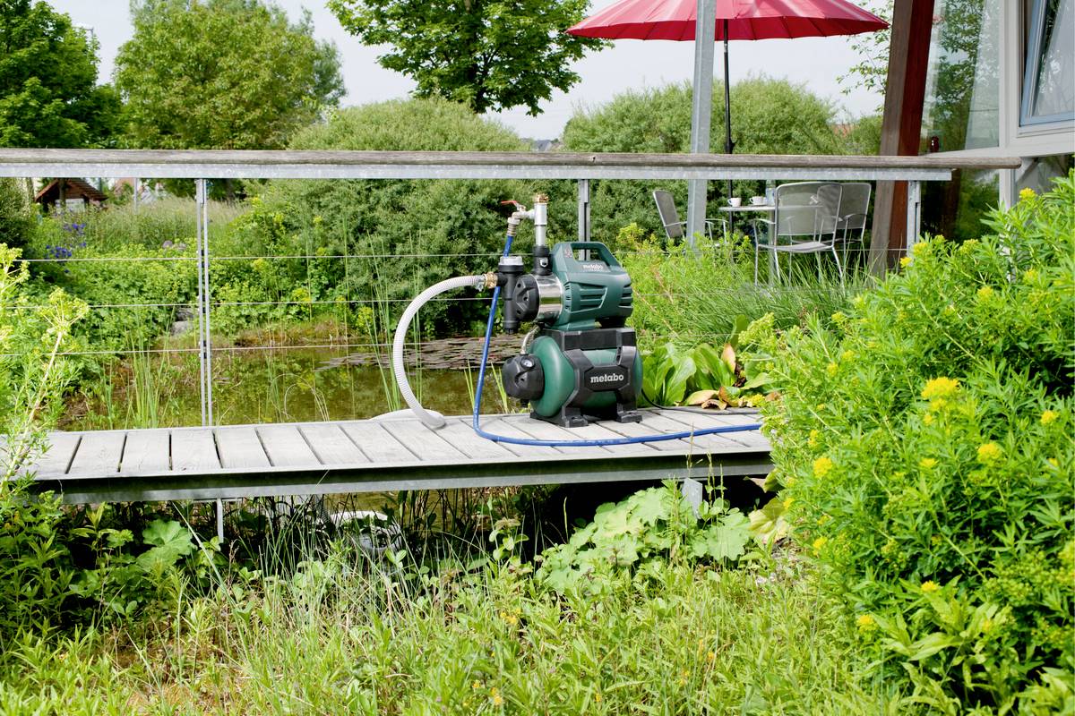 Een groene waterpomp staat op een houten steiger voor een vijver, omgeven door weelderige planten en struiken in de tuin.