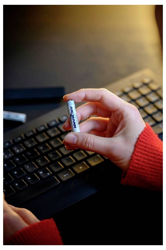 Een hand houdt een Panasonic AA-batterij boven een zwart computertoetsenbord, met een zichtbare rode trui mouw.