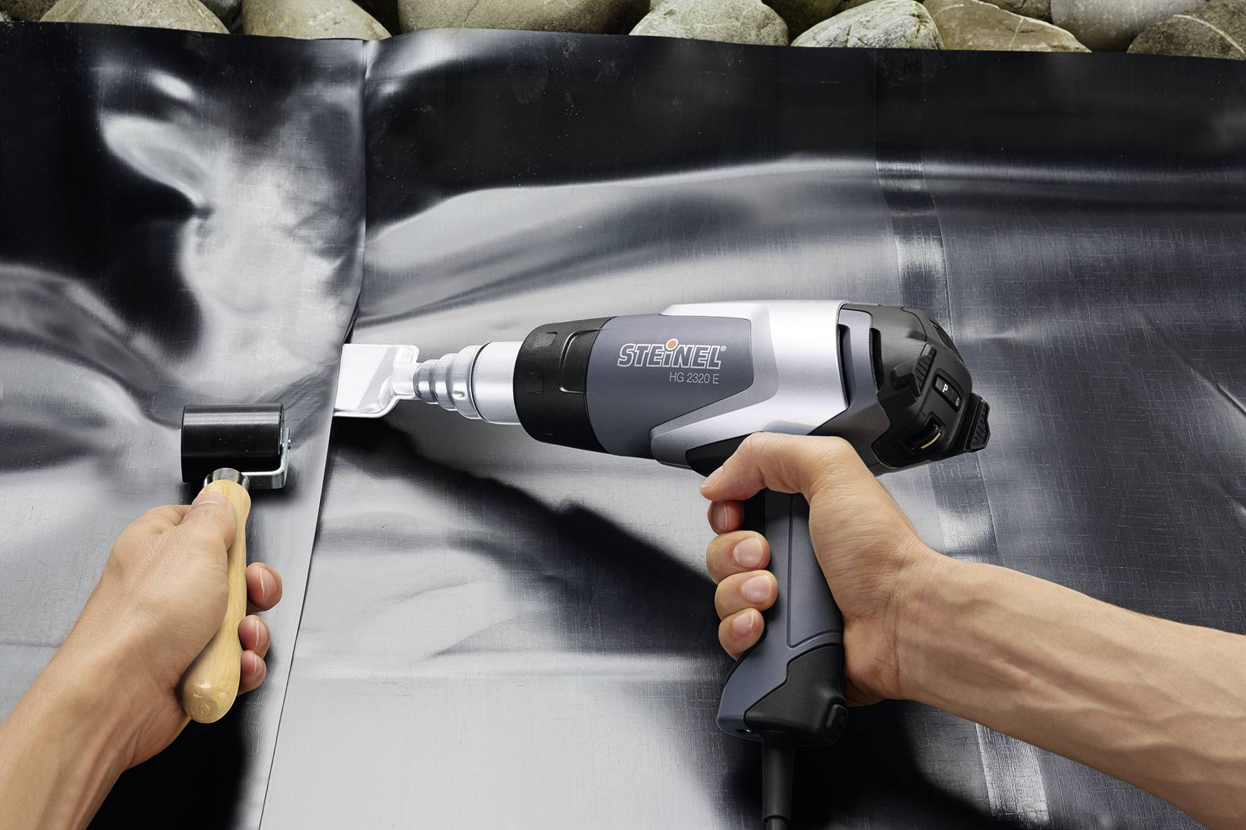 Een persoon bewerkt een zwarte folie met een heteluchtpistool en een aandrukrol. Op de achtergrond zijn stenen zichtbaar.