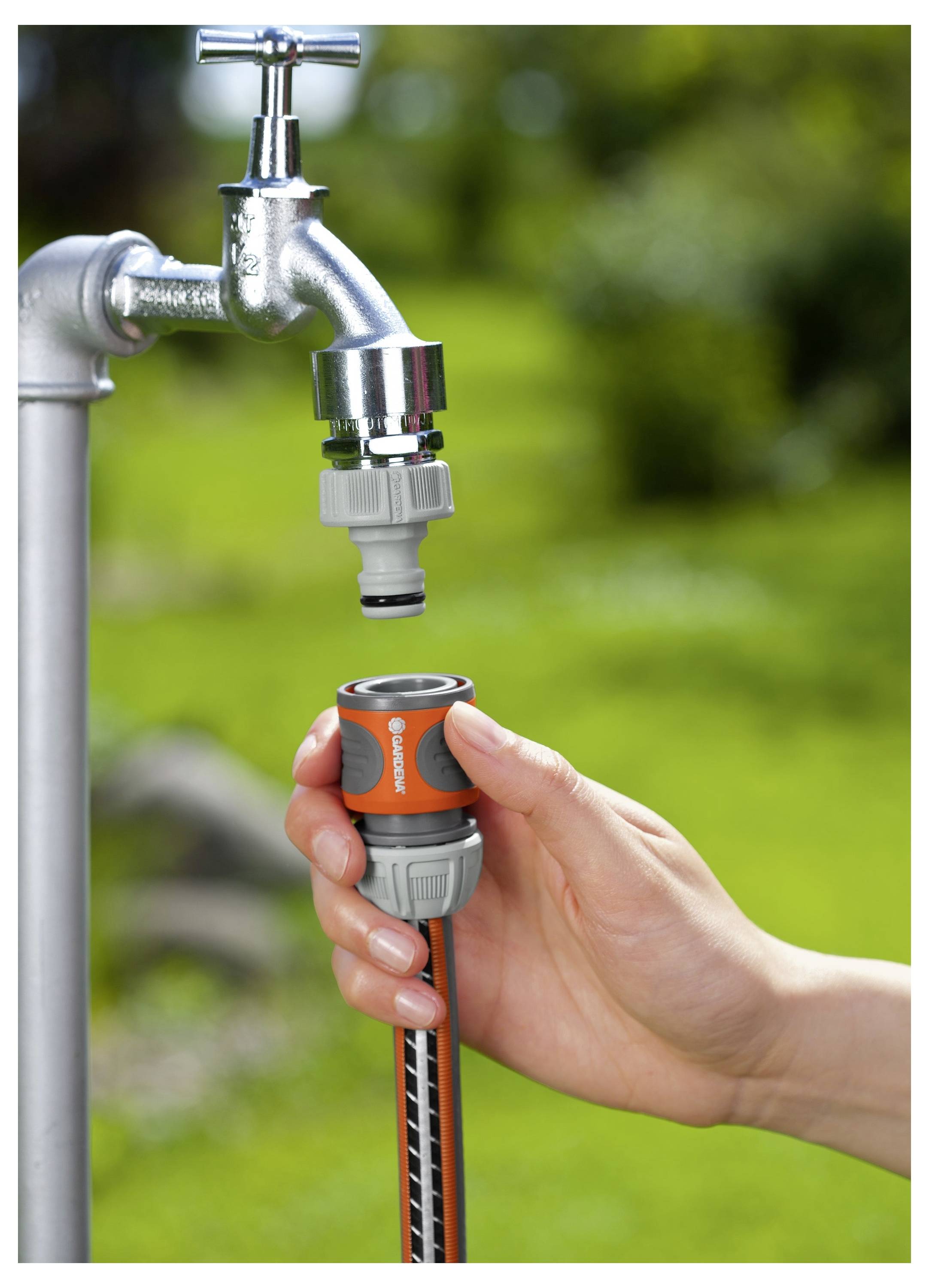 Een hand verbindt een tuinslang met een metalen buitenkraan met behulp van een oranje en grijze adapter, met een wazig groene achtergrond.