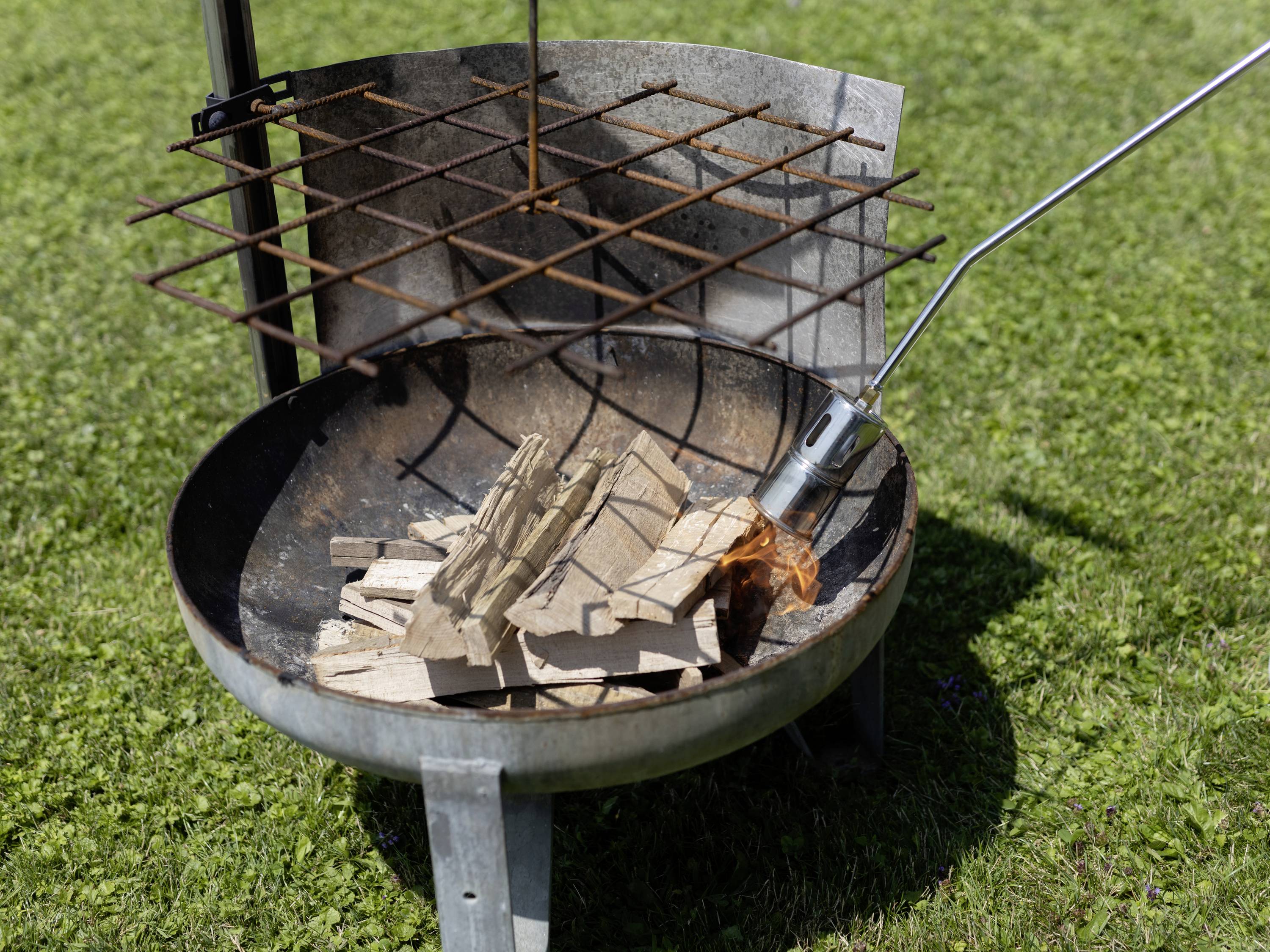 Een metalen vuurplaats met een rooster erbovenop, waar houtblokken worden aangestoken met een vlammenwerper. Gras op de achtergrond.