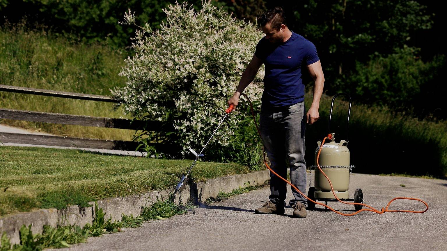Een persoon gebruikt een brander om onkruid langs de rand van een geplaveid pad te verwijderen. Op de achtergrond is een tuin zichtbaar.