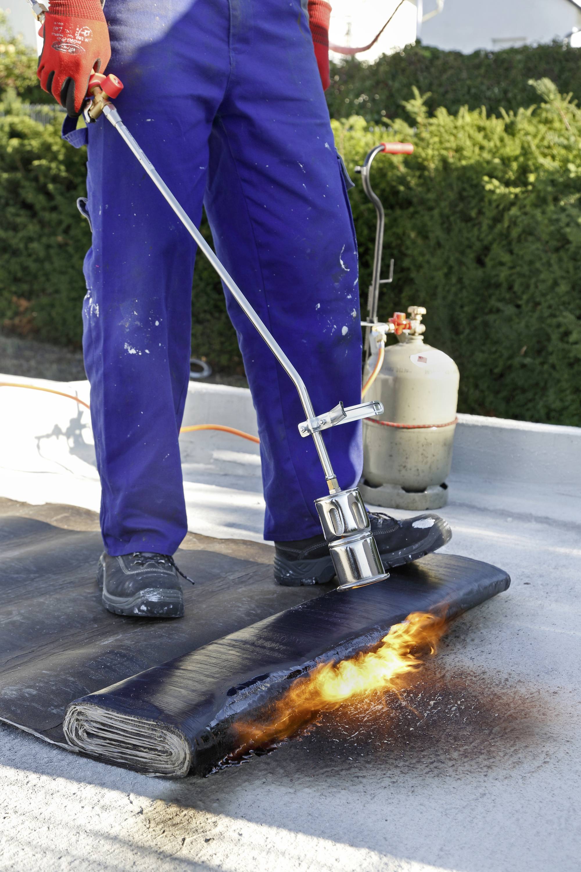 Een persoon draagt beschermende kleding en gebruikt een gasbrander om dakbedekking te lassen. Op de grond ontsteekt de laag.