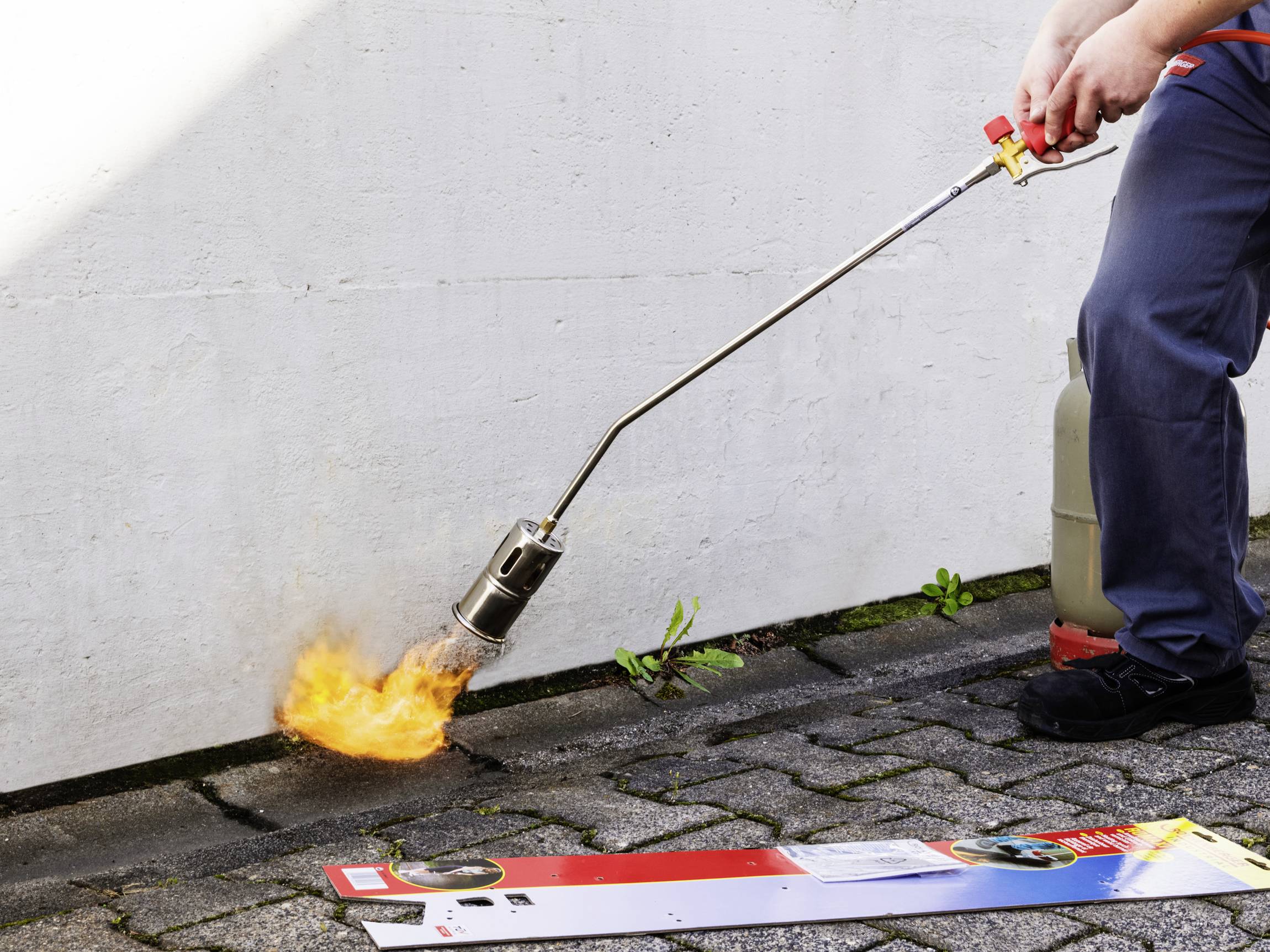 Een persoon gebruikt een onkruidbrander om onkruid tussen de bestratingstegels te verwijderen. Een jerrycan staat ernaast.