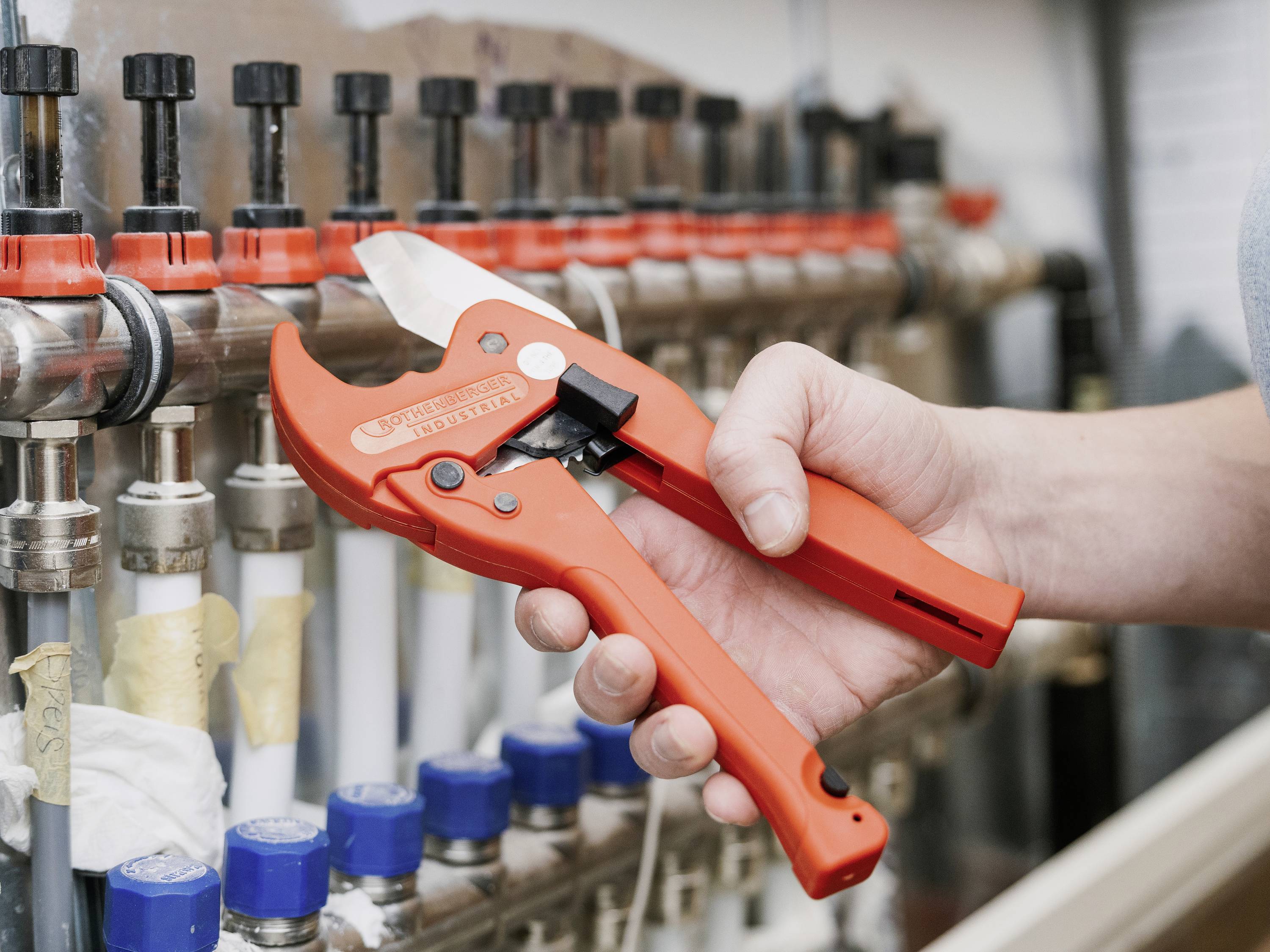 Een hand houdt een rode pijpsnijder vast voor een rij buizen en afsluiters die deel uitmaken van een verwarmingssysteem.