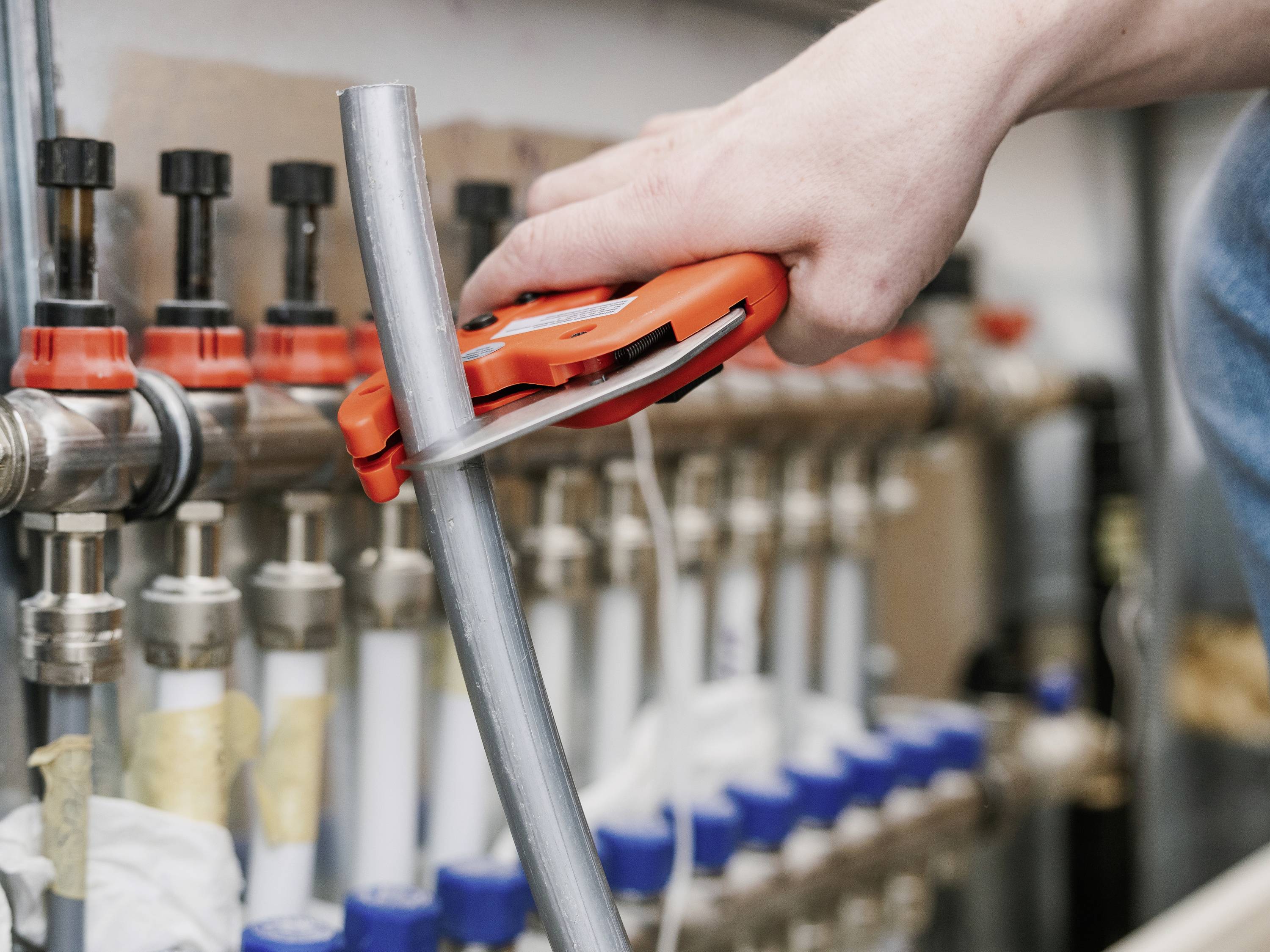 Een hand bedient een gereedschap dat een metalen buis doorsnijdt. Op de achtergrond zijn aansluitingen en kleppen zichtbaar.