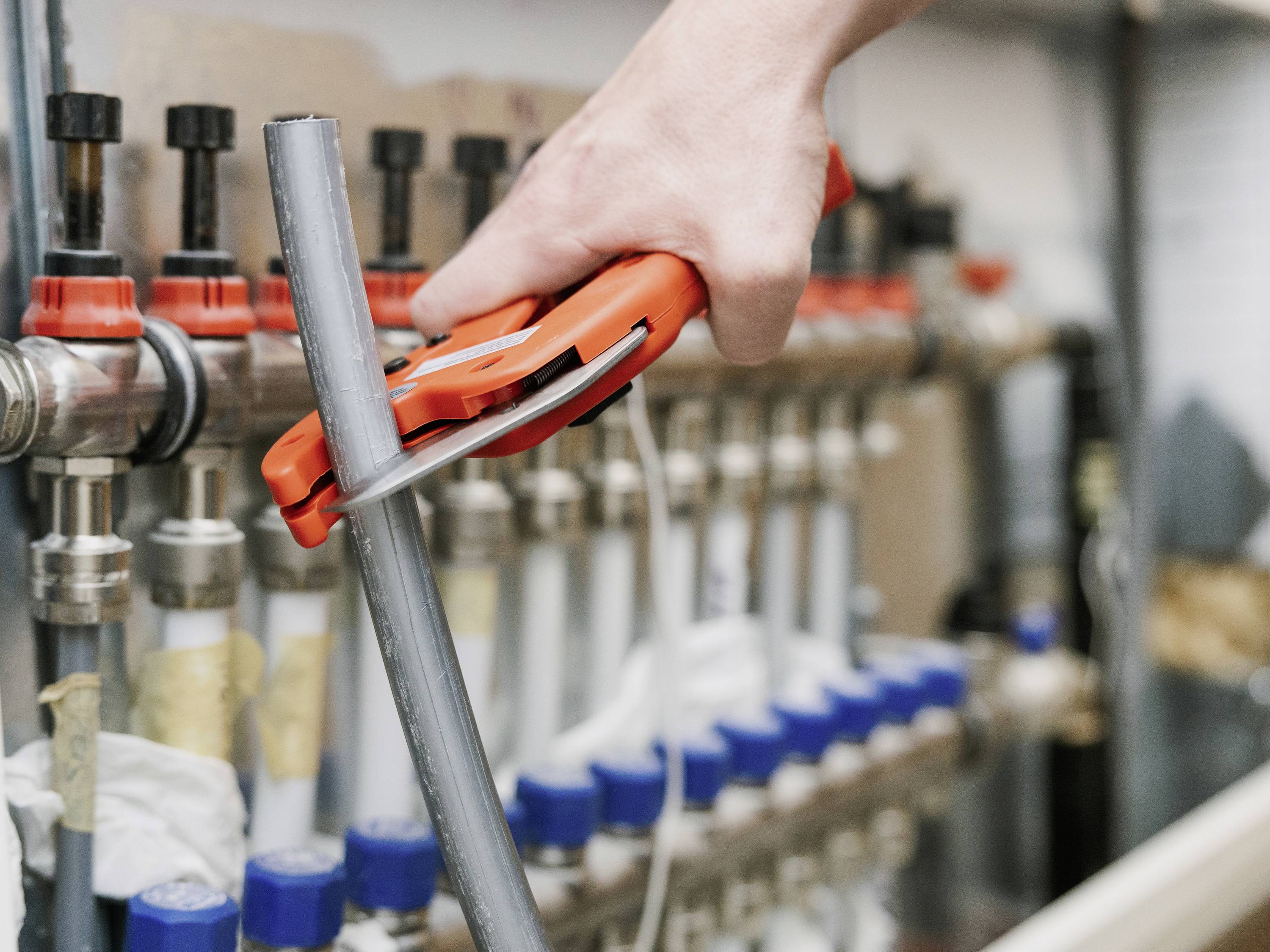 Een persoon snijdt een metalen buis met een buizensnijder. Op de achtergrond zijn verschillende buizen en afsluiters te zien aan een verwarmingsinstallatie.