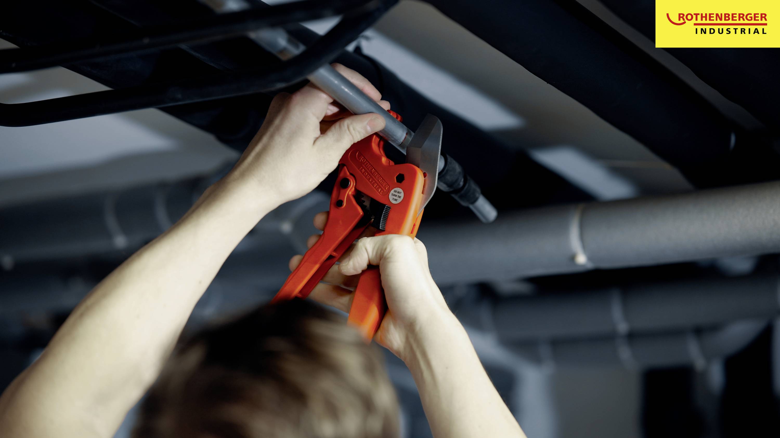 Twee handen bedienen een oranje pijptang die werkt aan een grijs buizensysteem. Op de achtergrond een industriële omgeving.