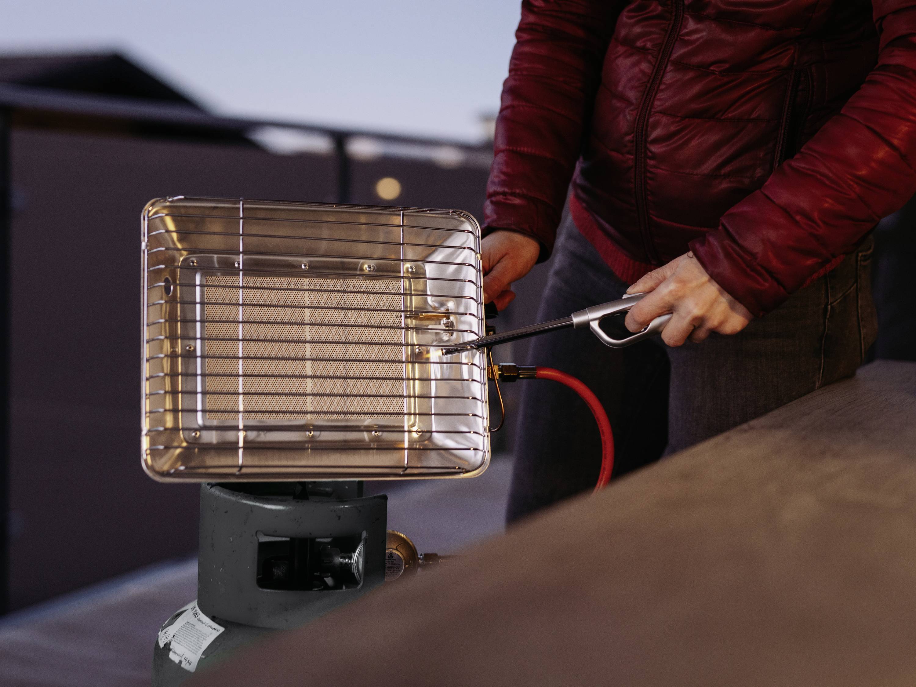 Een persoon in een rode jas steekt met een aansteker een gasgestookte verwarmer aan op een terras of balkon.