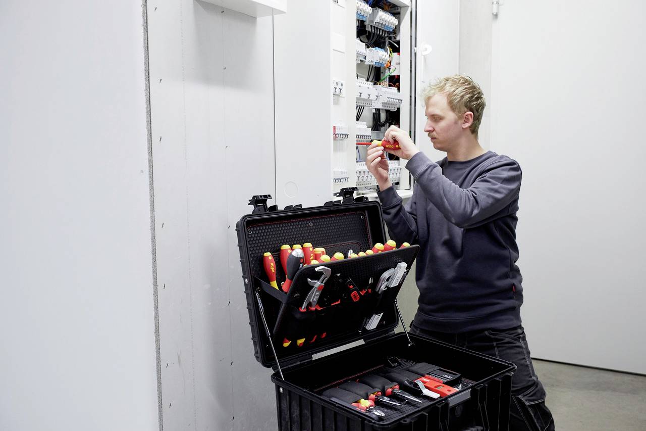 Een man werkt aan een open schakelkast, met een schroevendraaier in zijn hand. Een gereedschapskoffer met gereedschappen staat naast hem.