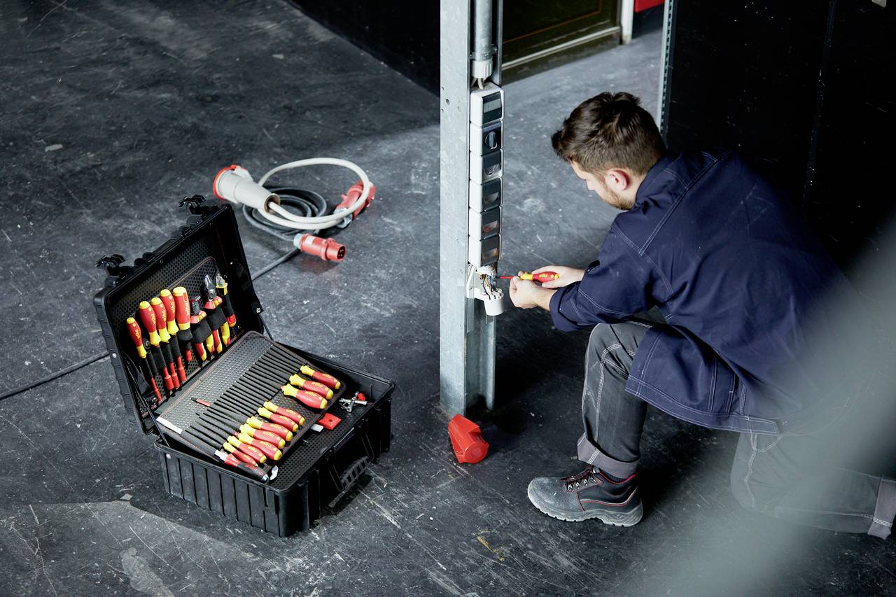 Een persoon werkt aan een stopcontact in een industriële omgeving. Naast hem staat een open gereedschapskist met schroevendraaiers op de grond.