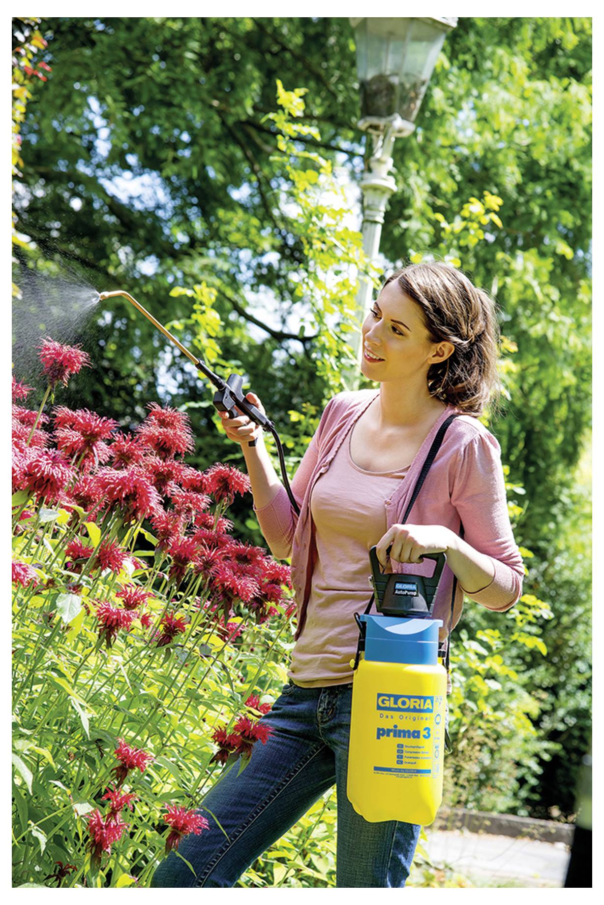 Een persoon gebruikt een tuinspuit om rode bloemen in een zonnig park te begieten. Ze houden een geel en blauw sproeier met 'Gloria'-branding vast.