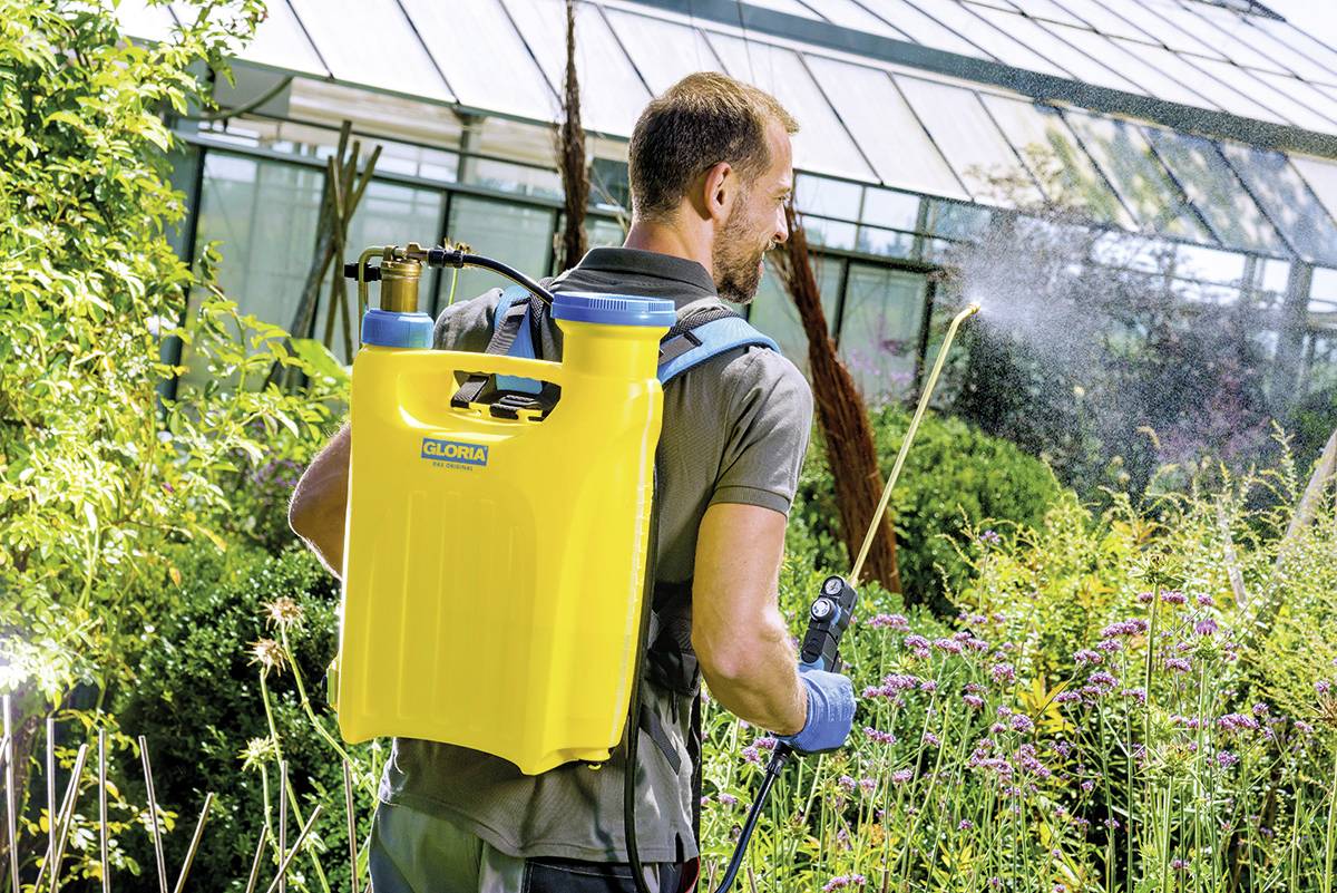 Een man met een gele rugzaksproeier bewatert planten in de tuin. Op de achtergrond een kas.