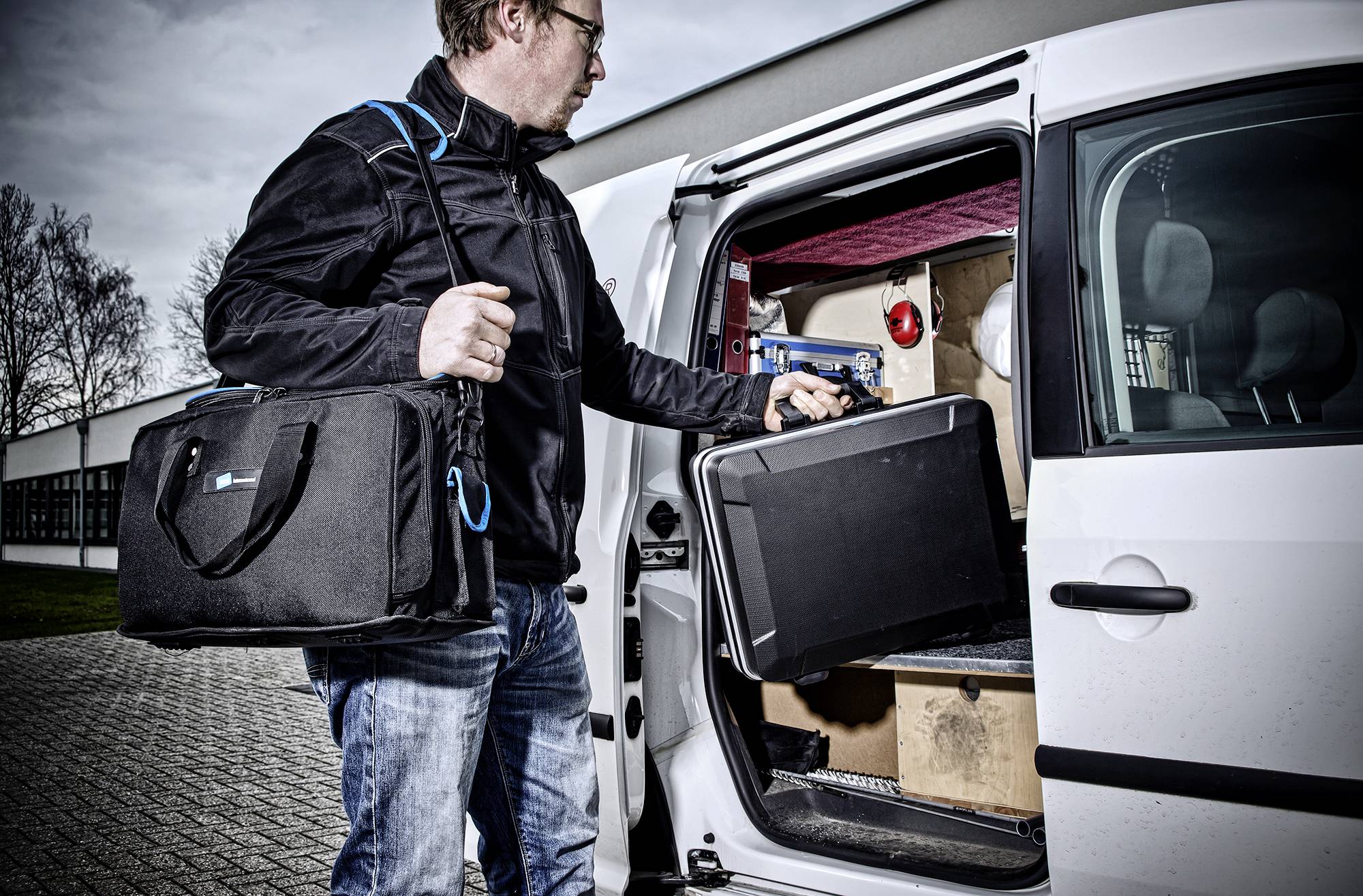 Een persoon draagt een zwarte tas en haalt een zwarte koffer uit een witte bestelwagen. Op de achtergrond zijn dozen gestapeld.