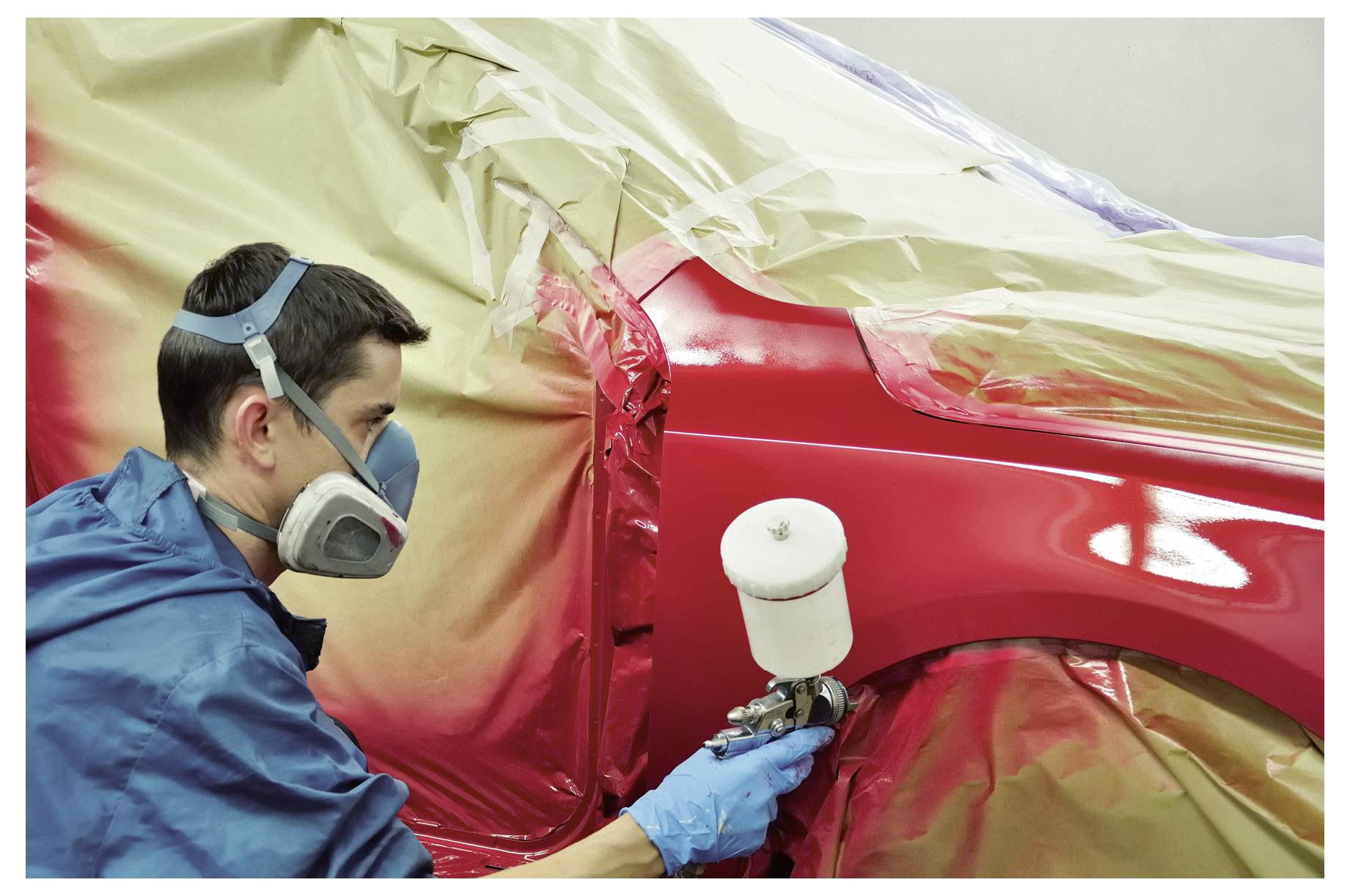 Een technicus besproeit een rood autobordeel met spuitbus in een karosseriewerkplaats, waarbij de rest van het voertuig met beschermend plastic en tape is afgekit.
