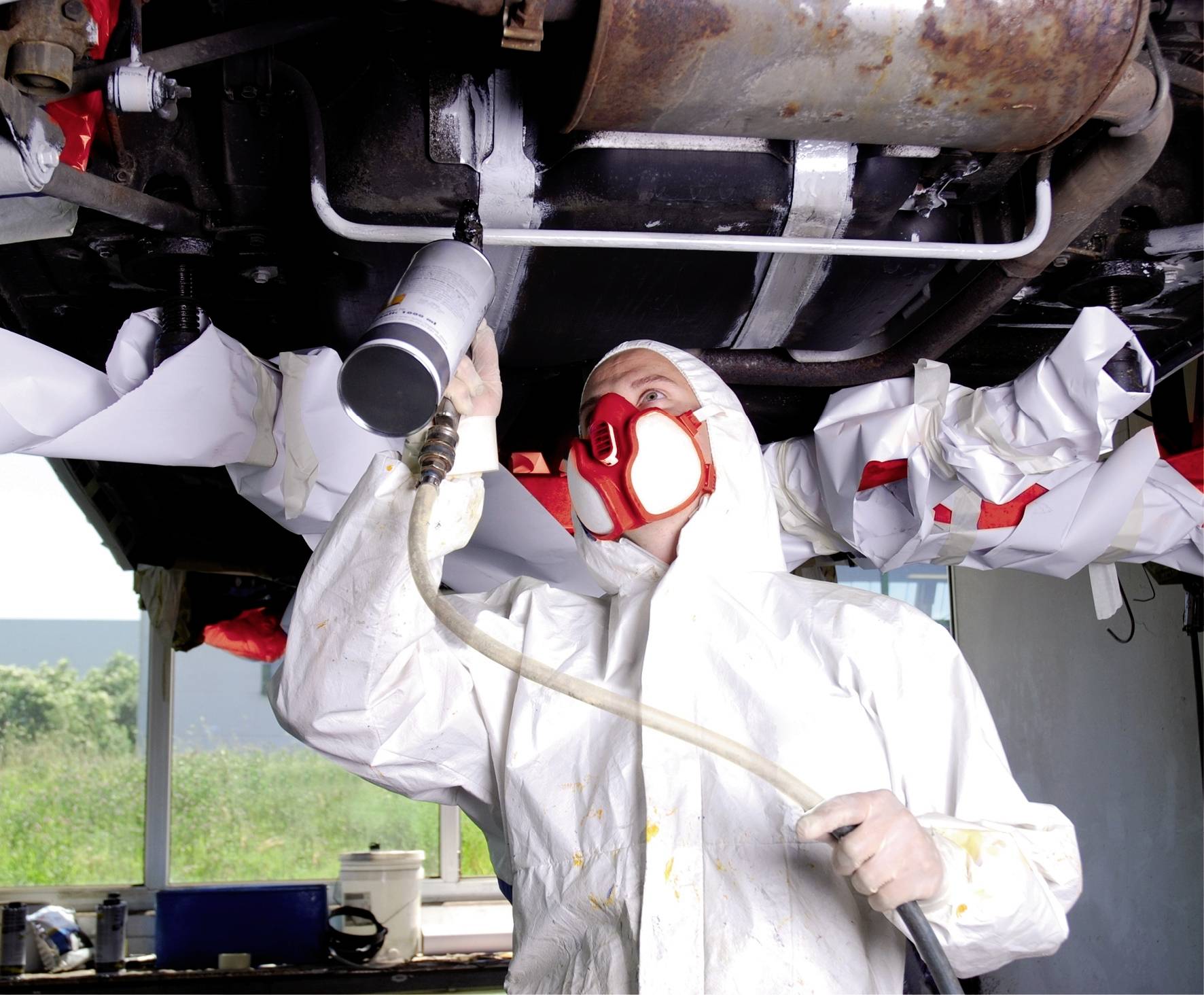 Een persoon in beschermende kleding spuit de onderkant van een auto in een werkplaats. Op de achtergrond zijn gereedschappen en groene landschappen zichtbaar.