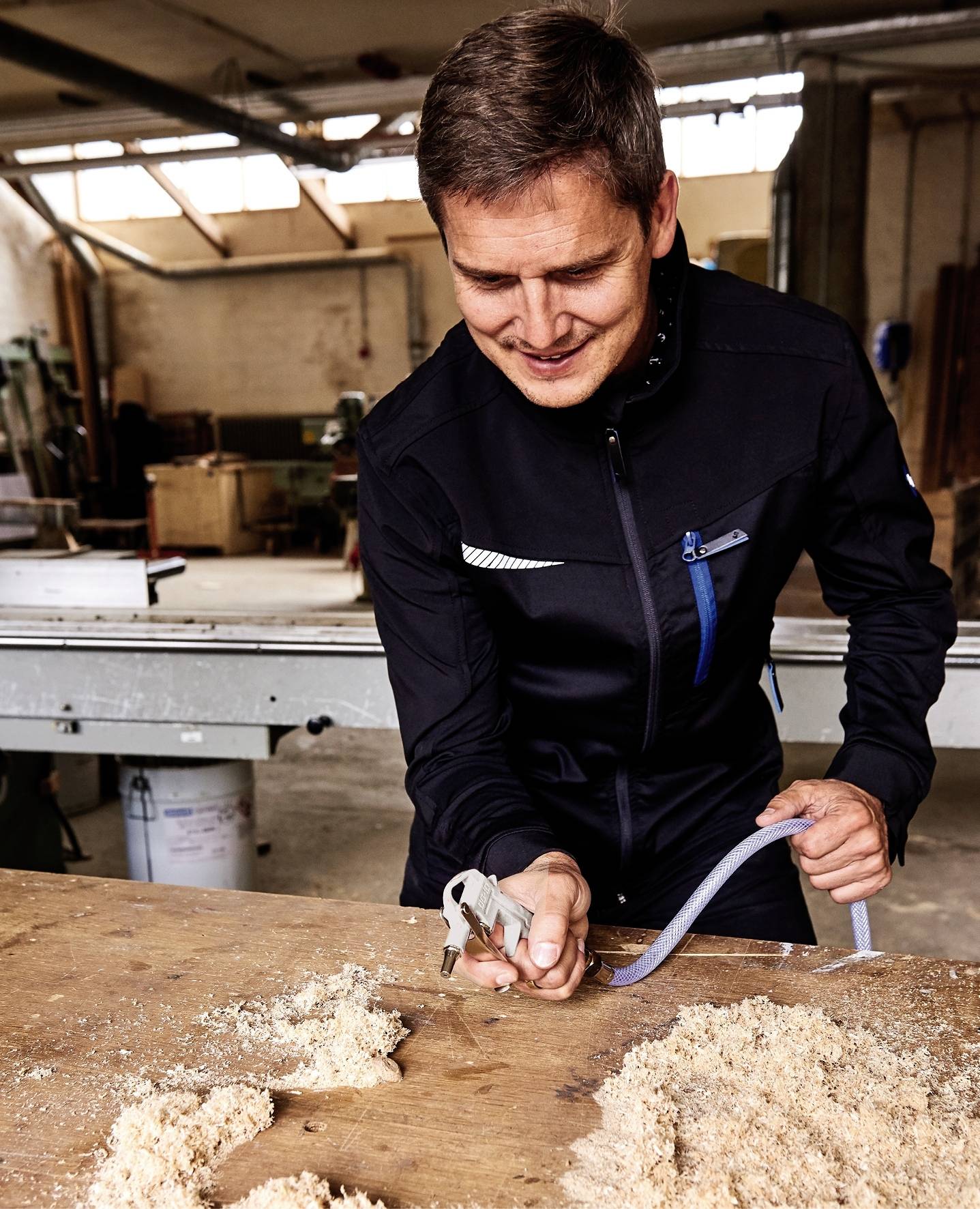Een man in een werkplaats glimlacht terwijl hij met een gereedschap werkt in houtkrullen. Op de achtergrond zijn machines en ramen zichtbaar.