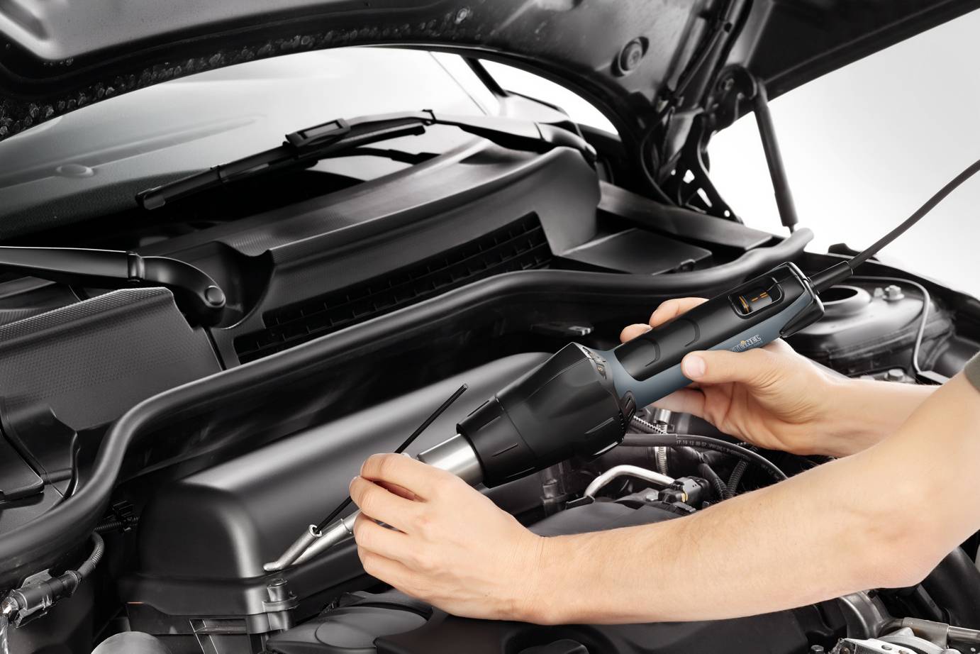 Een persoon repareert een automotor met een elektrisch gereedschap. De afbeelding toont gedetailleerd de motorkap en het gereedschap.