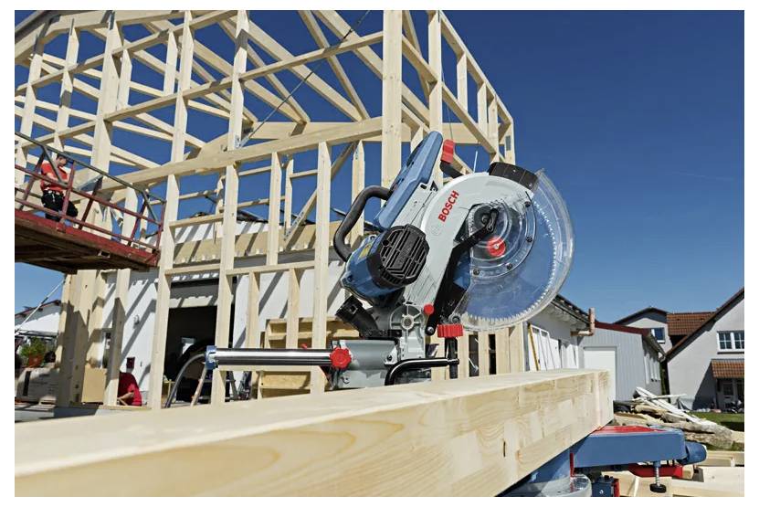 Een cirkelzaag snijdt een houtbalk door op een bouwplaats van een huisconstructie onder een heldere blauwe lucht.
