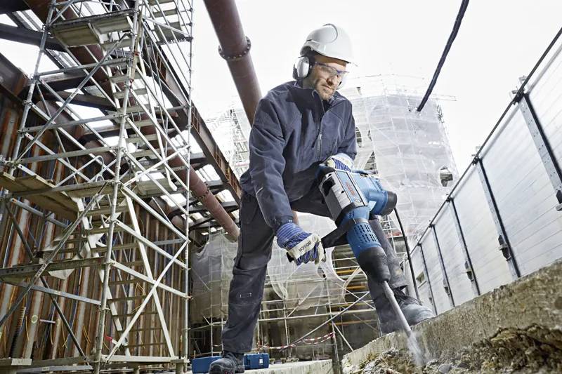 Een bouwvakker in veiligheidsuitrusting gebruikt een jackhammer om de grond te breken op een bouwterrein, met steiger en gebouwen zichtbaar op de achtergrond.