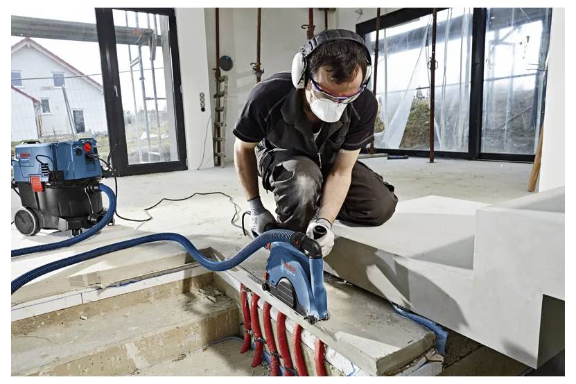 Een werknemer gebruikt een elektrisch gereedschap om beton in een deels gebouwd gebouw te snijden, draagt persoonlijke beschermingsmiddelen en knielt naast trappen.