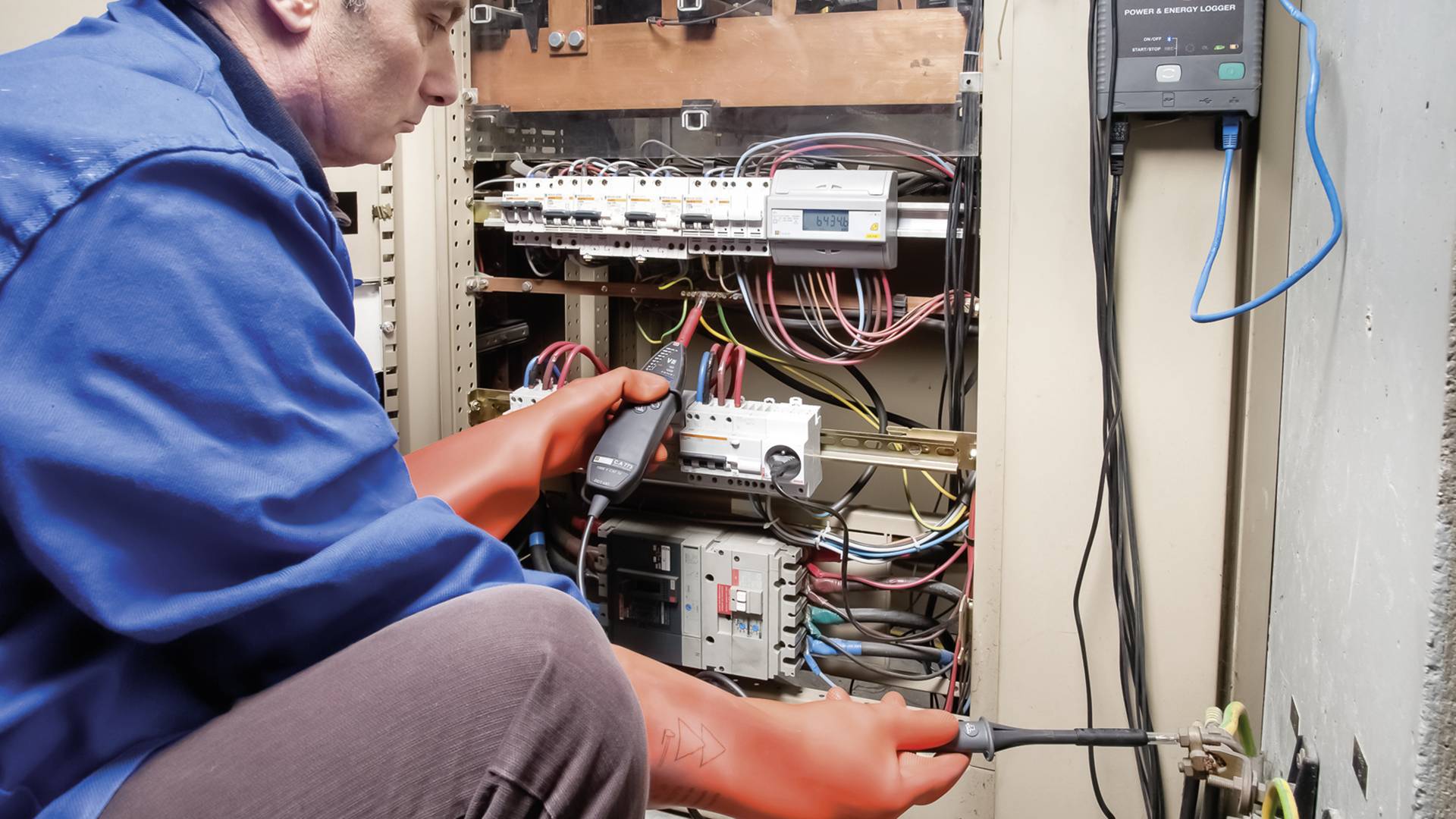 Een elektricien in blauwe werkkleding en oranje handschoenen controleert met een apparaat een open schakelkast vol kabels.