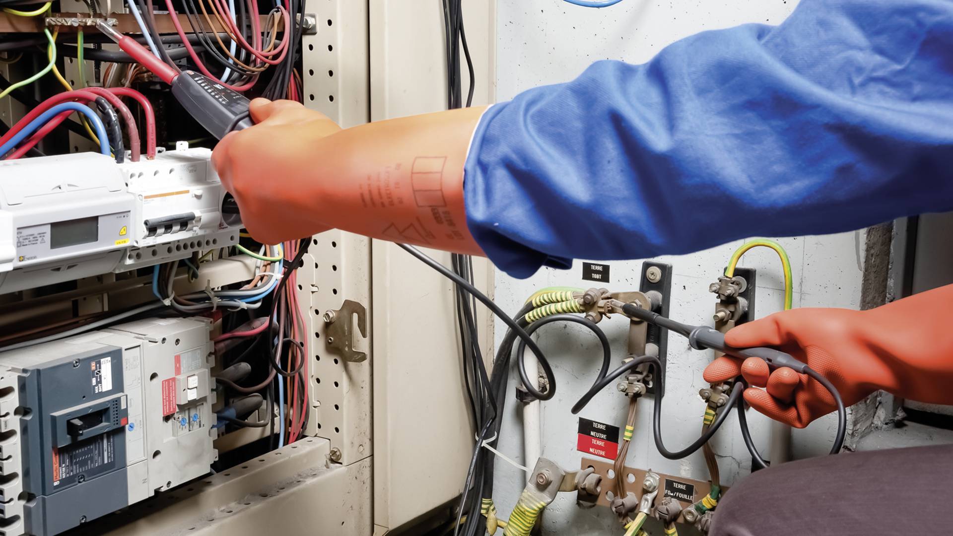 Een persoon in beschermende kleding werkt aan een schakelkast, waarbij verbindingen en kabels worden gecontroleerd met een elektronisch apparaat.