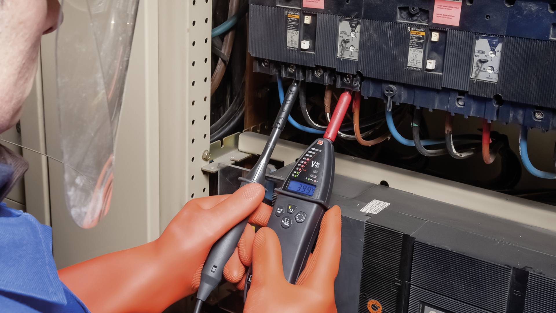 Een persoon met veiligheidsbril en -handschoenen meet spanning aan een open zekeringkast met een digitaal multimeter.