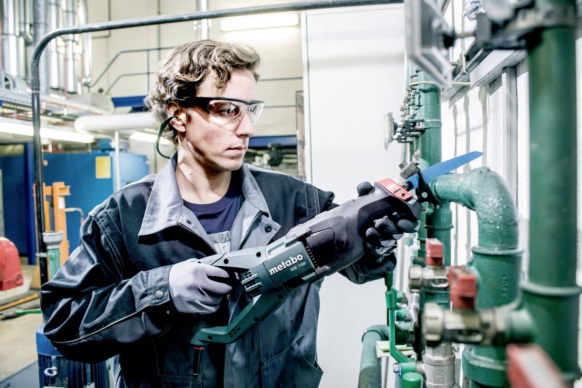 Een arbeider met veiligheidsbril en handschoenen gebruikt een Metabo-reciprozaag om aan buizen te werken in een industriële omgeving.