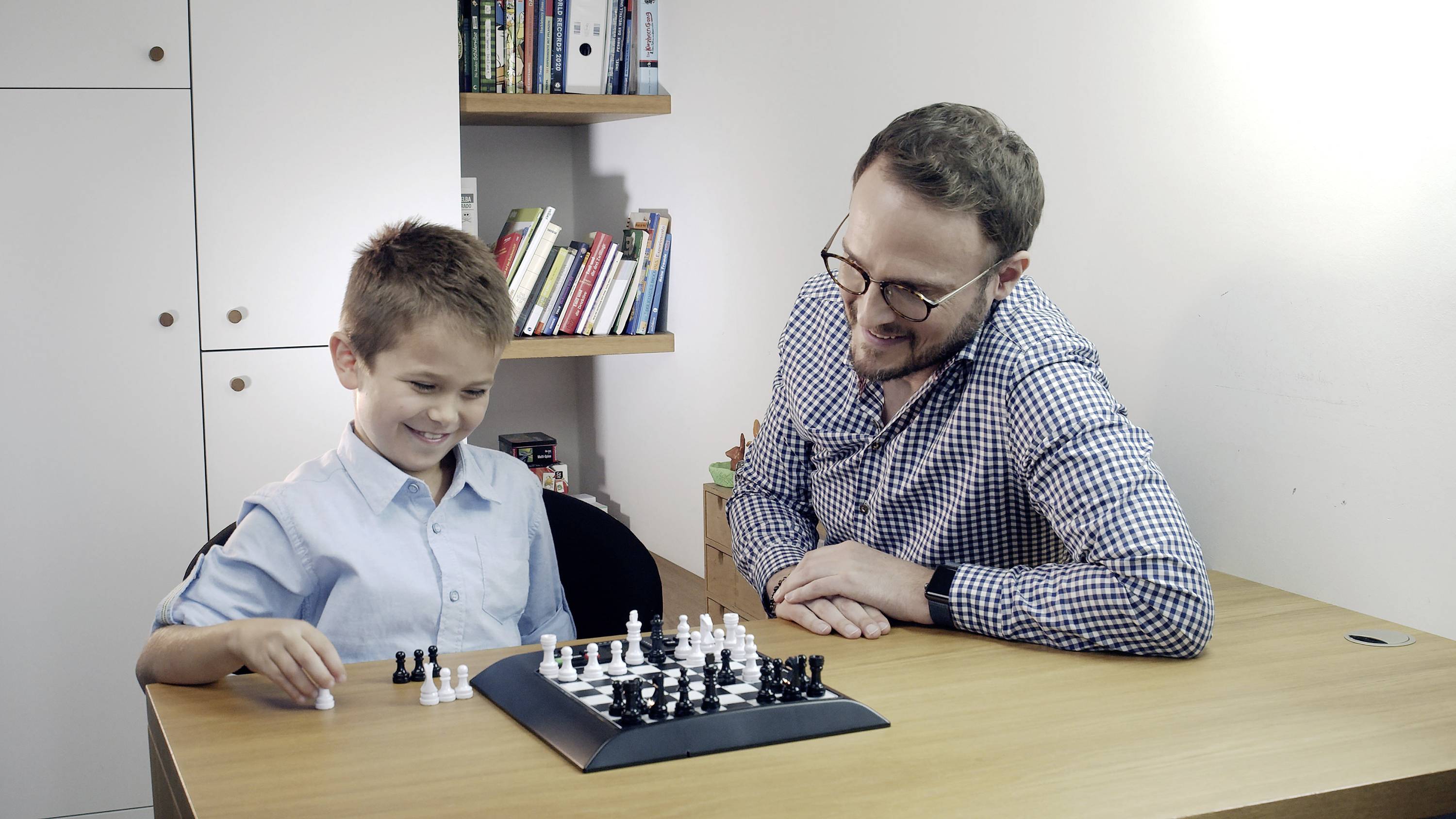 Een man en een jongen spelen schaak aan een houten tafel. De jongen is blij met zijn zet. Op de achtergrond staat een boekenkast.