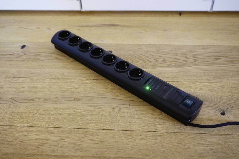 Een zwarte stekkerdoos met zeven aansluitingen ligt op een houten vloer. Een groene controlelamp is ingeschakeld.