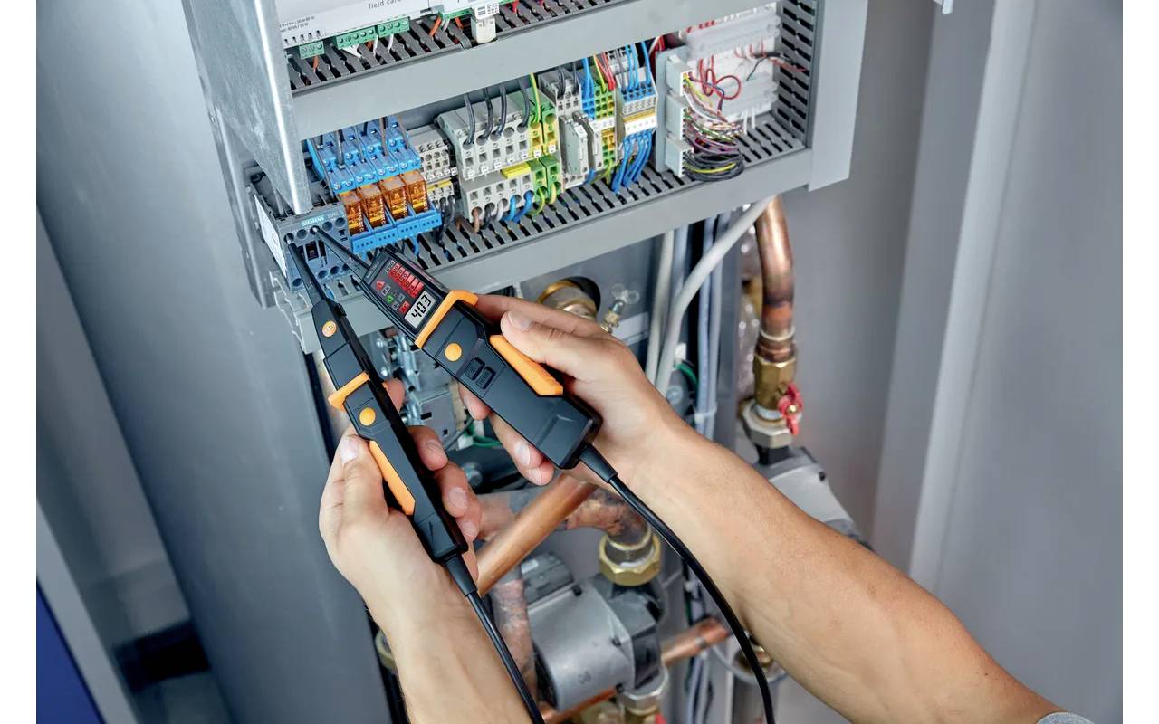 Een technicus gebruikt een tang om elektrische draden in een schakelkast te inspecteren. Op de achtergrond zijn verschillende kabels zichtbaar.