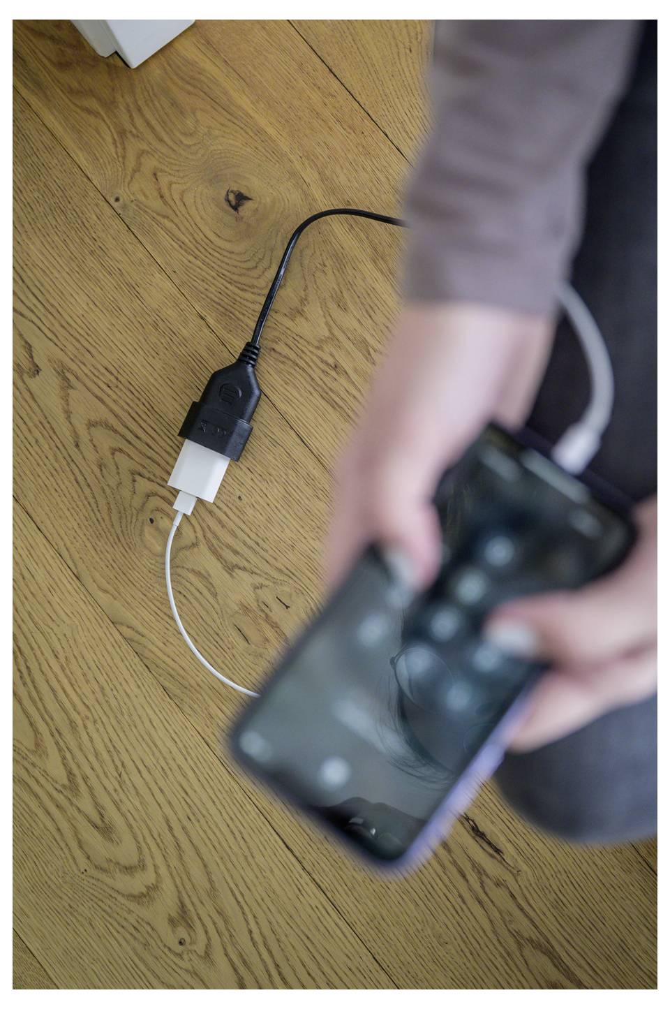 Een persoon die een smartphone vasthoudt terwijl deze via een kabel die is verbonden met een wandadapter op een houten vloer oplaadt.