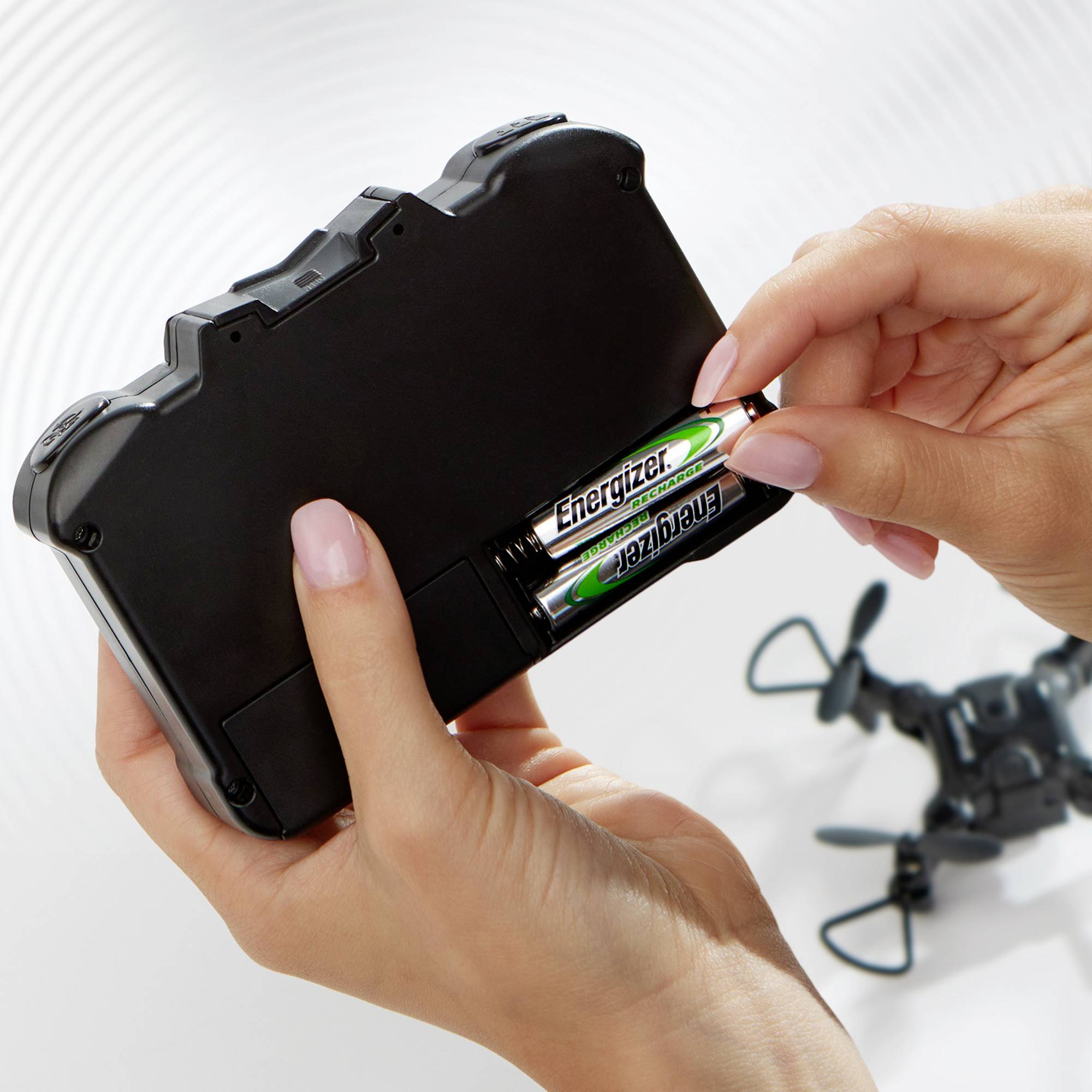 Een persoon plaatst twee Energizer-batterijen in een zwart elektronisch apparaat. Op de achtergrond is een klein speelgoedvliegtuigje te zien.